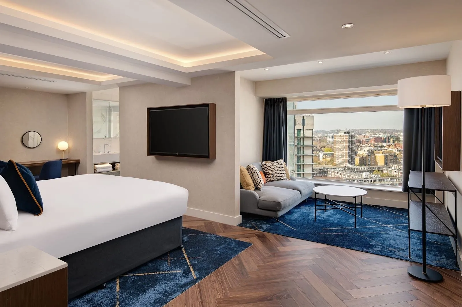 A large room at Hilton London Metropole with a king bed, herringbone floors under navy rugs, and a grey sofa and round coffee table in a picture window looking out over tower blocks
