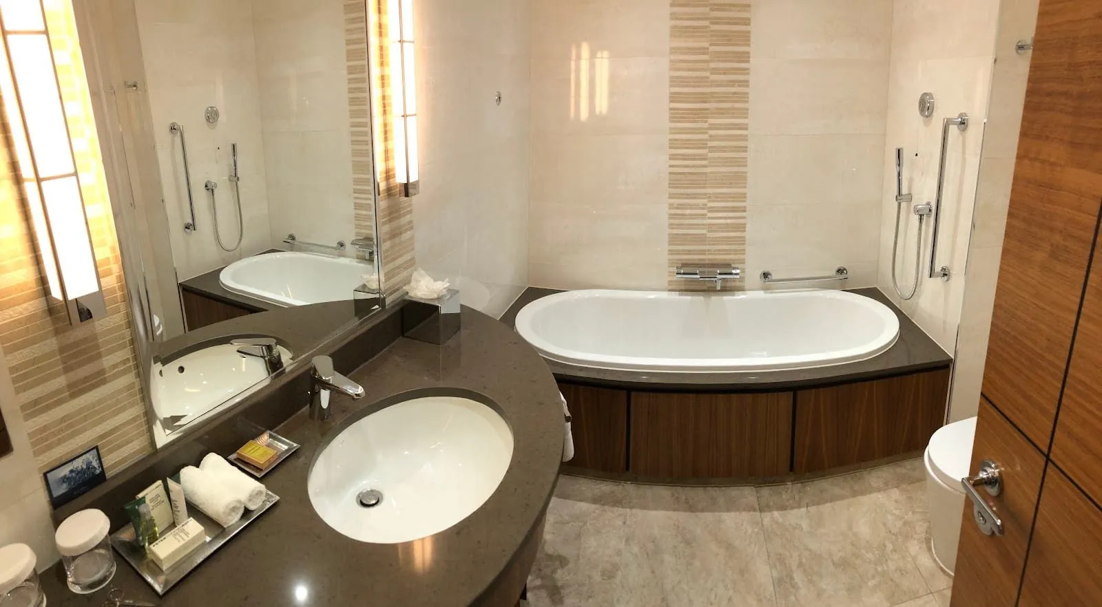 A bathroom at Hilton London Paddington with a deep oval bath set in walnut panelling, a hand shower in a glass-screened corner, grab rails and a curved stone vanity