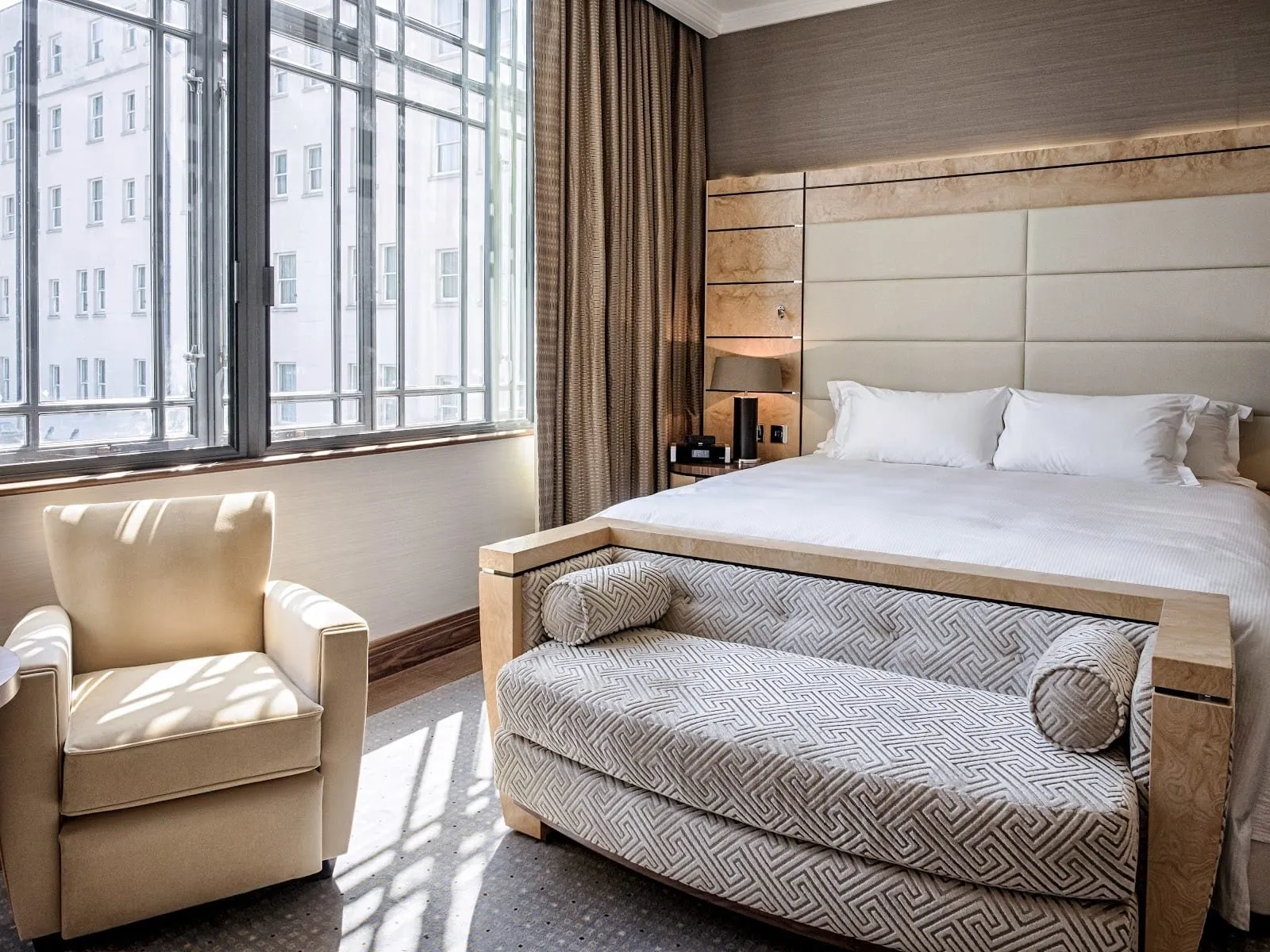 A double room at Hilton London Paddington with tall steel-framed windows, a cream armchair, a pale burr-wood headboard and an upholstered bench across the foot of the bed