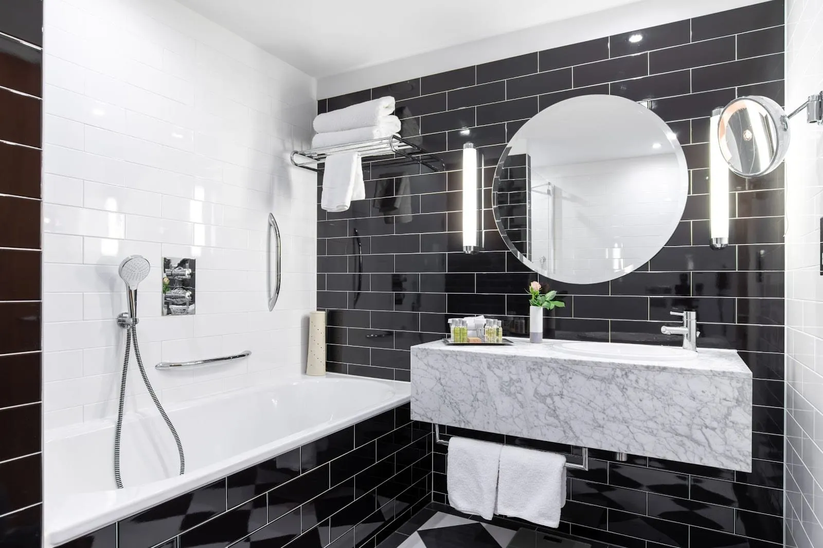 A bathroom at the Hilton London Wembley with black and white metro tiles, a bath with a hand shower, a round mirror and a white marble basin unit