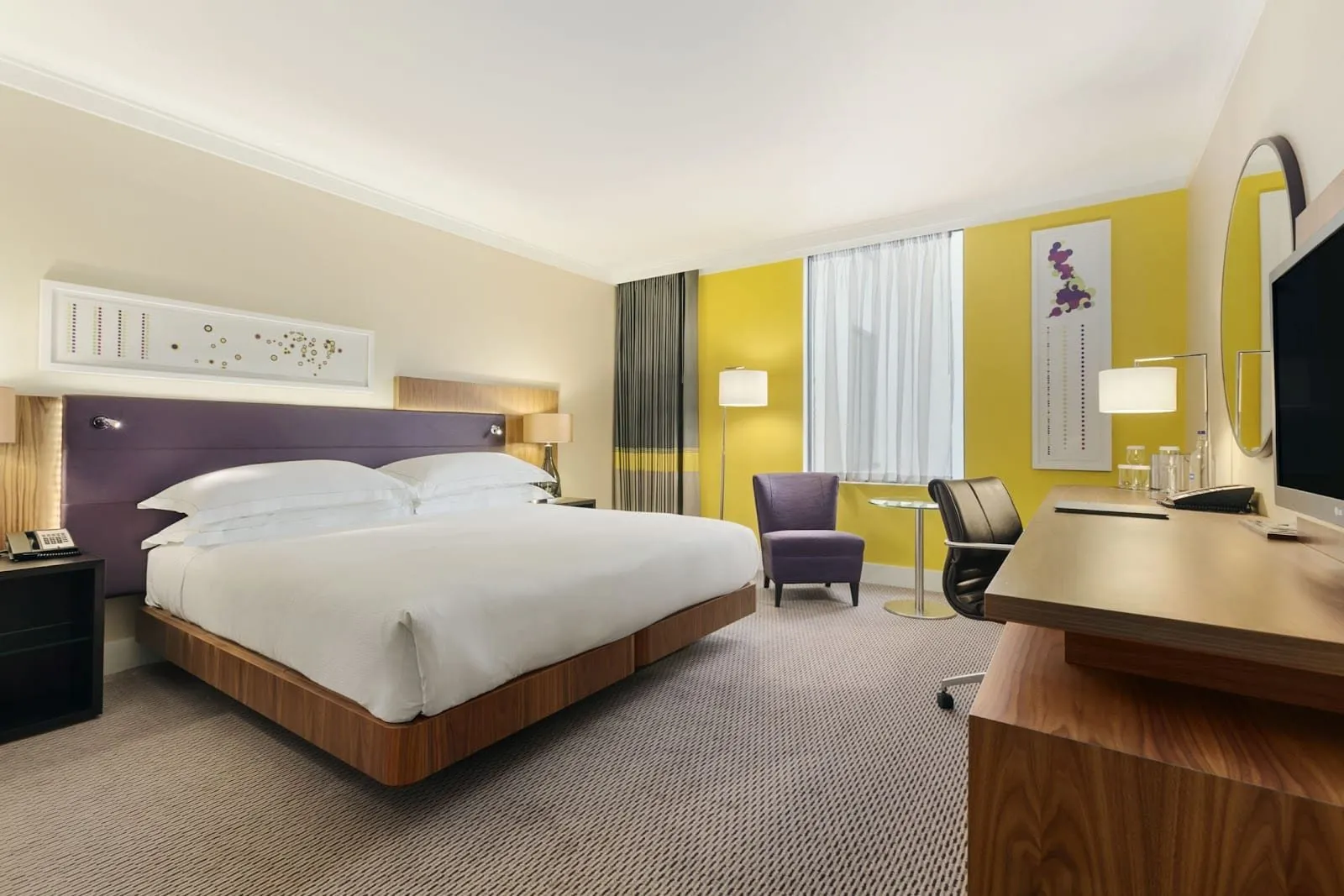 A room at the Hilton London Wembley with a king-size bed against a purple padded headboard, a yellow feature wall by the window, a purple armchair and a walnut desk