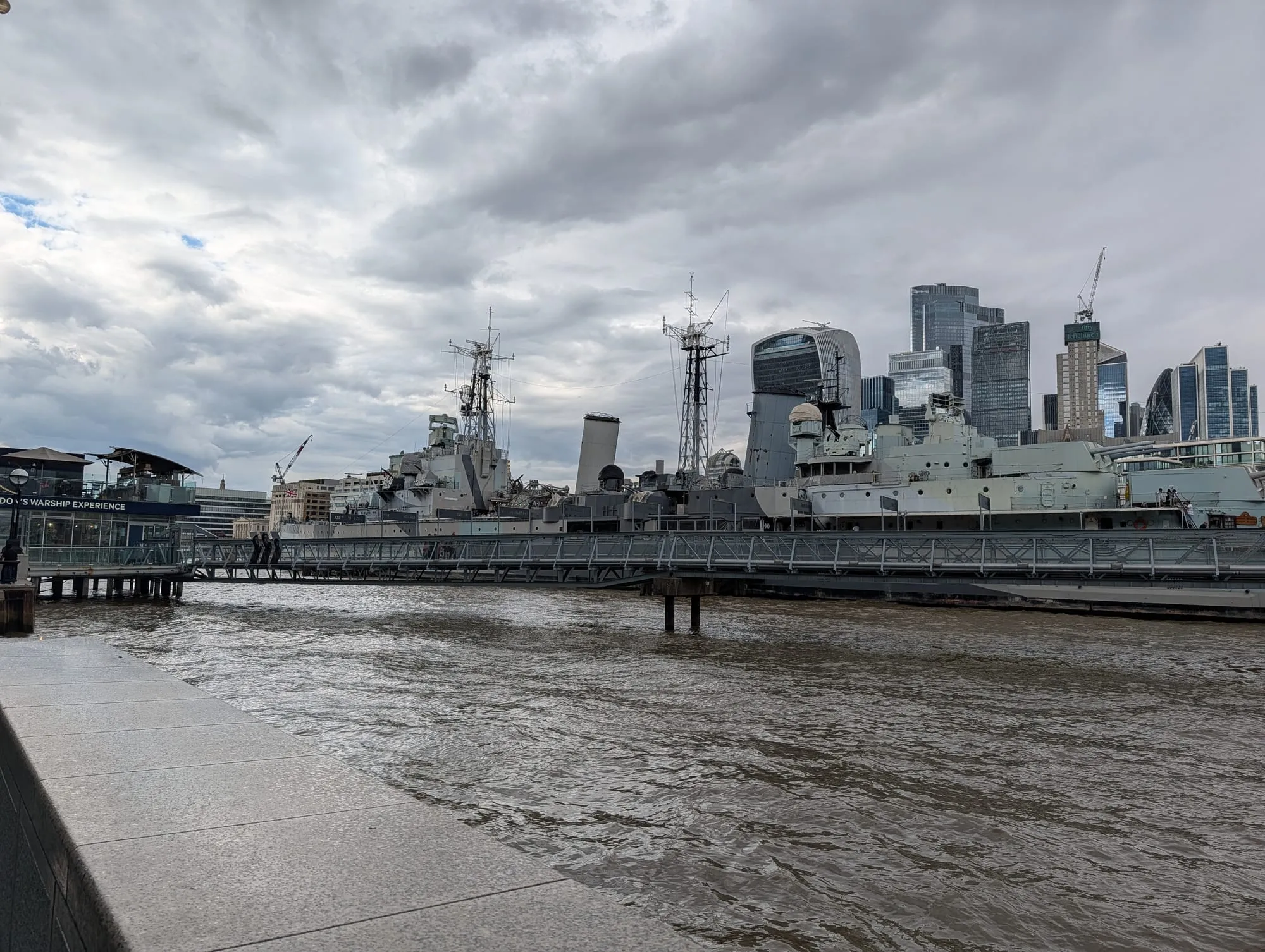 HMS Belfast moored on the Thames with the City of London's towers behind it, the Walkie-Talkie and the Gherkin on the skyline