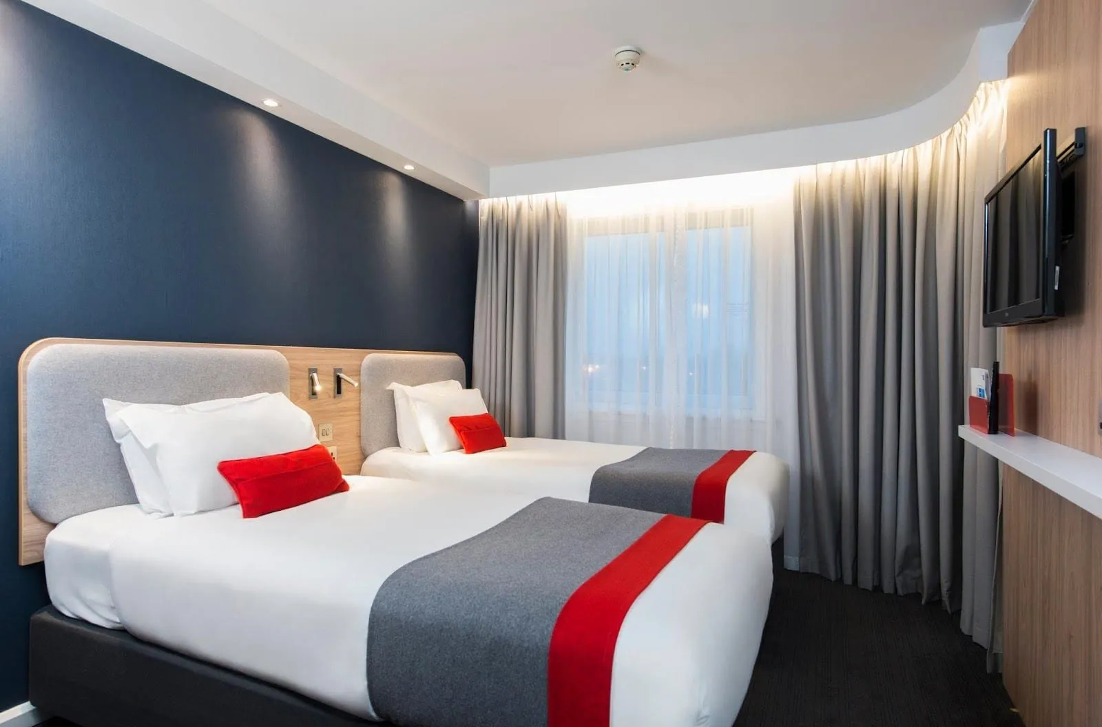 A twin room at Holiday Inn Express London – Greenwich with two single beds under grey padded headboards, red cushions and grey-and-red runners, a navy wall, a lit curtain pelmet and a wall-mounted TV