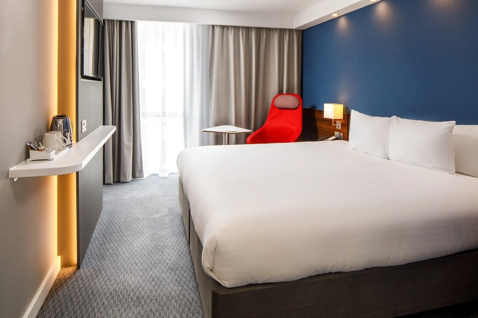 A double room at Holiday Inn Express London – ExCeL with a blue feature wall, a red chair and a kettle shelf