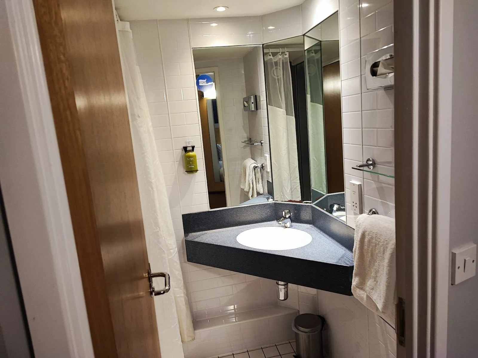 A small bathroom at Holiday Inn Express London – Limehouse with a corner basin on a navy counter, angled mirrors, white metro tiles and a shower behind a white curtain
