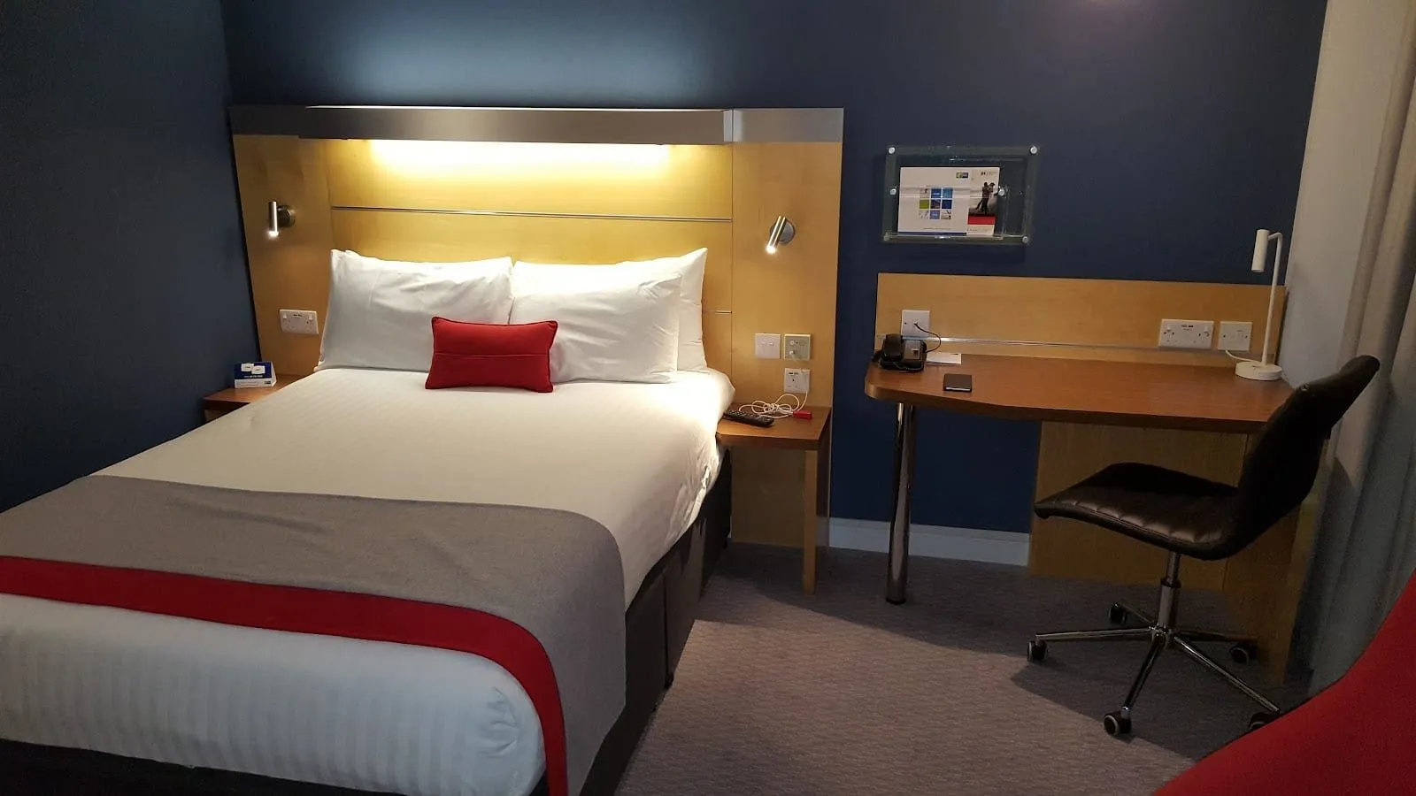 A double room at Holiday Inn Express London – Limehouse with a pale wood headboard lit from above, a red cushion, navy walls and a wood desk with a black office chair
