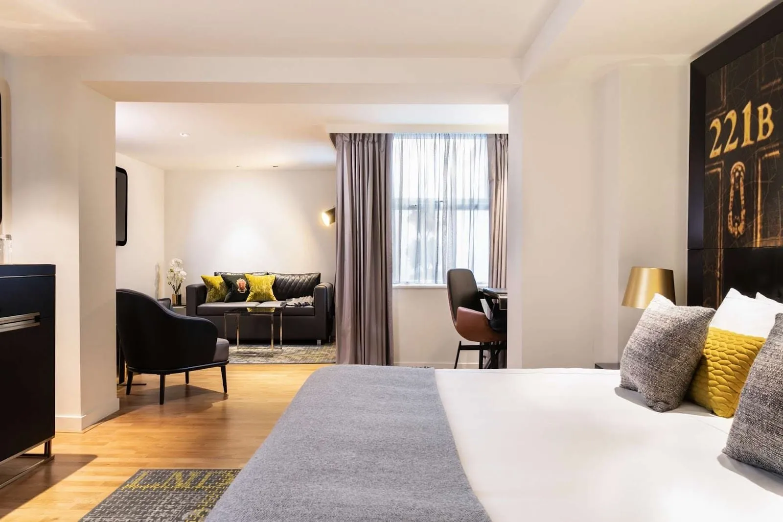 A suite at Holmes Hotel London with the bed's black headboard printed with a gold 221B door and knocker, oak floors, and a sitting area with a leather sofa and yellow cushions through a wide opening