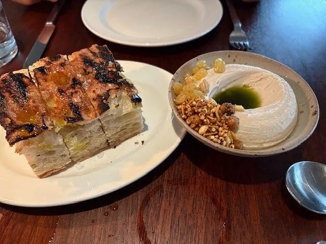 Charred squares of grilled focaccia on a white plate beside a bowl of whipped dip topped with green oil, crisp chickpeas and golden raisins