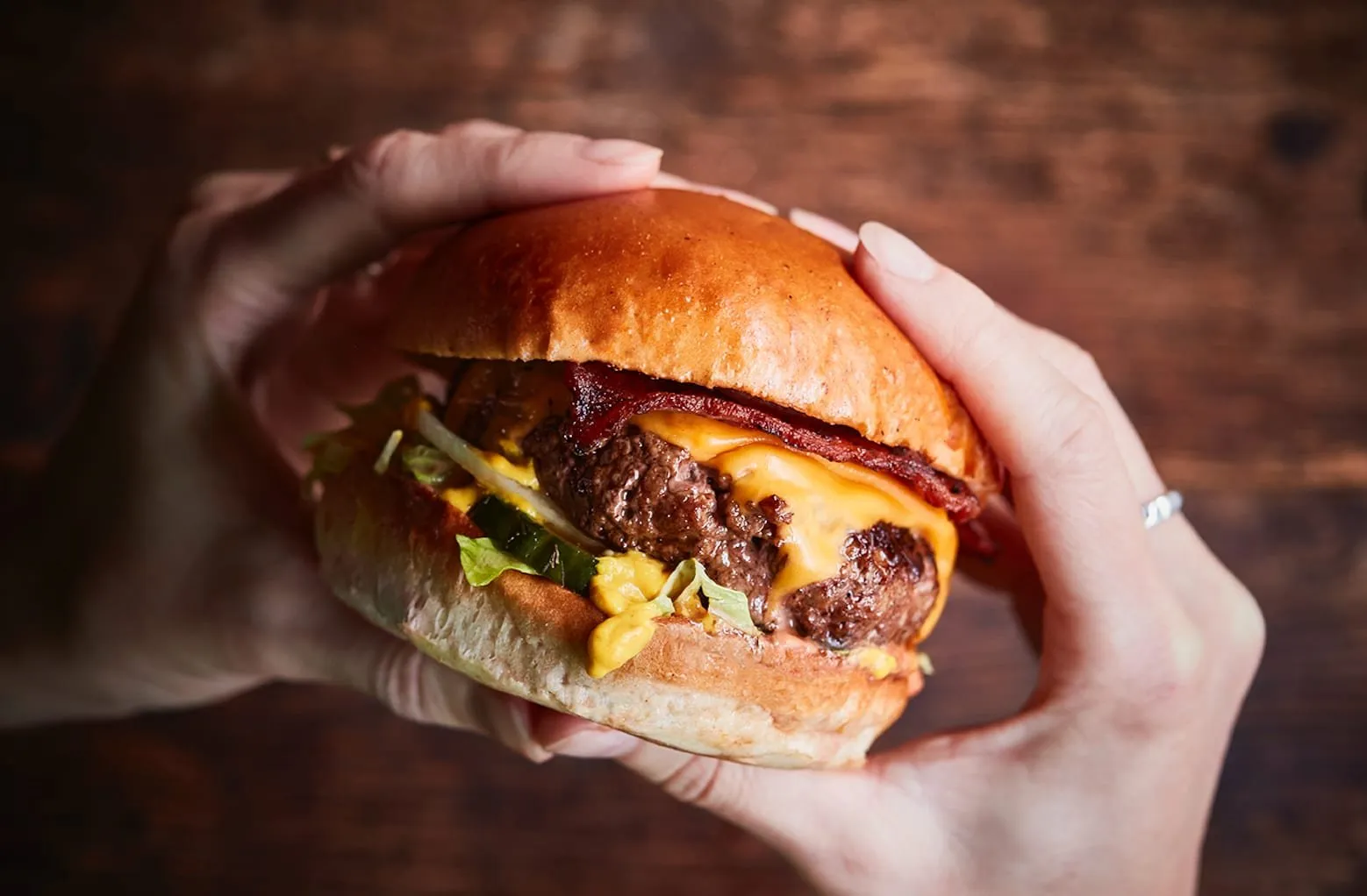 An Honest Burgers cheeseburger held in two hands, a thick beef patty under melted cheese and crisp bacon, with pickles, lettuce and mustard sauce in a glazed brioche bun