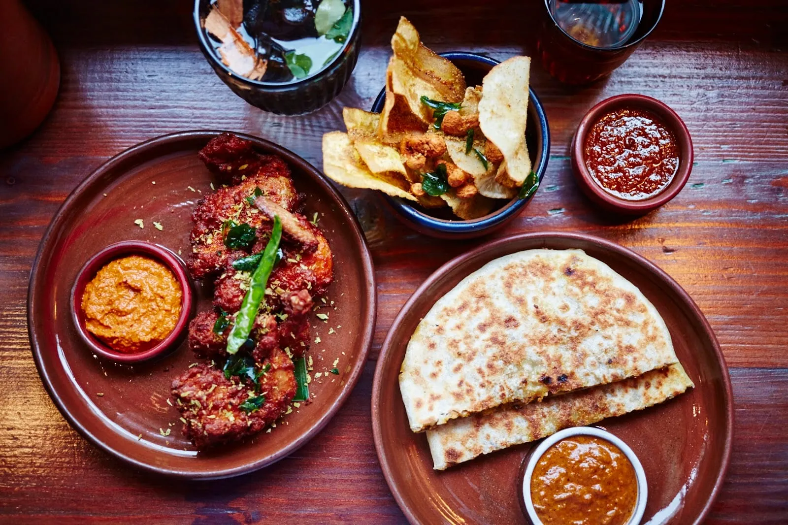 An overhead spread on a dark wooden table: a plate of red-spiced devilled meat with curry leaves and green chilli, a bowl of curry-leaf crisps, two clay pots of sambol, and a folded godhamba roti with a pot of curry sauce