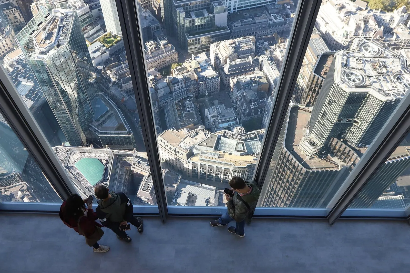 Three visitors at floor-to-ceiling windows looking straight down onto City rooftops, a glass-roofed atrium and an office tower with plant on its roof