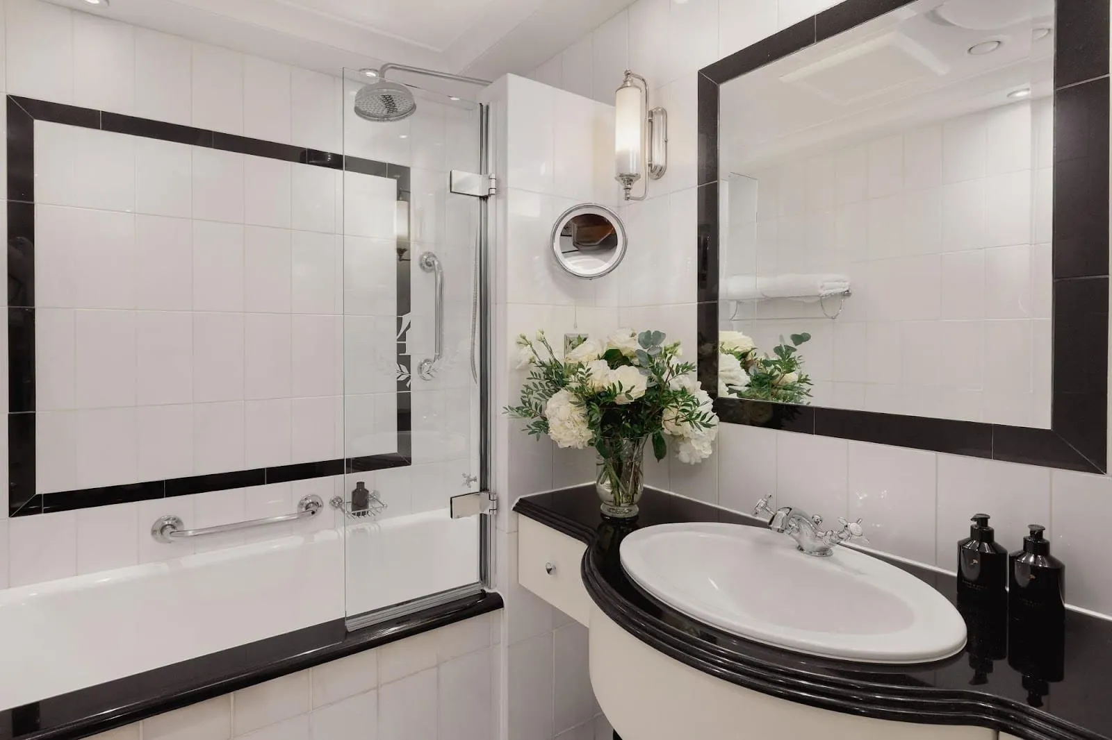 A bathroom at Hotel 41 with a bath and overhead rain shower behind a glass screen, white tiles framed in black, and a curved black-topped vanity with a vase of white flowers