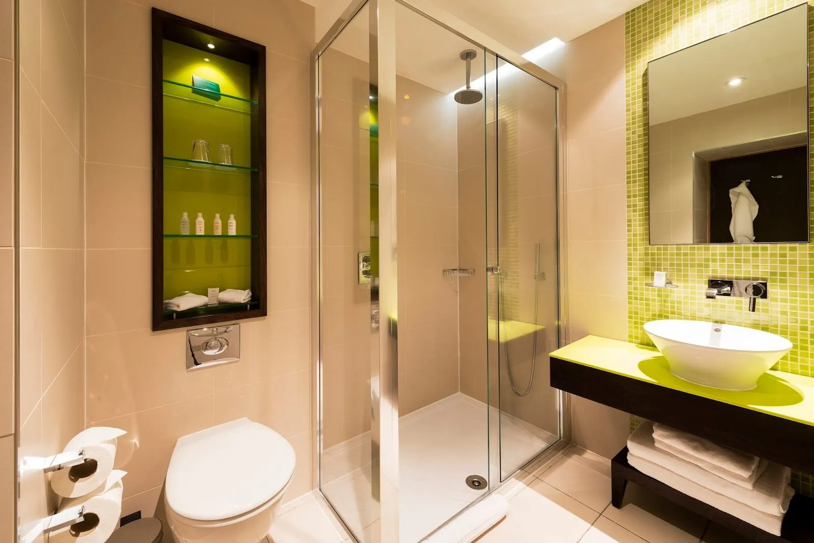 A bathroom at Hotel Indigo London Paddington with a glass shower cubicle under a rainfall head, a lime-green mosaic wall and a bowl basin on a lime counter