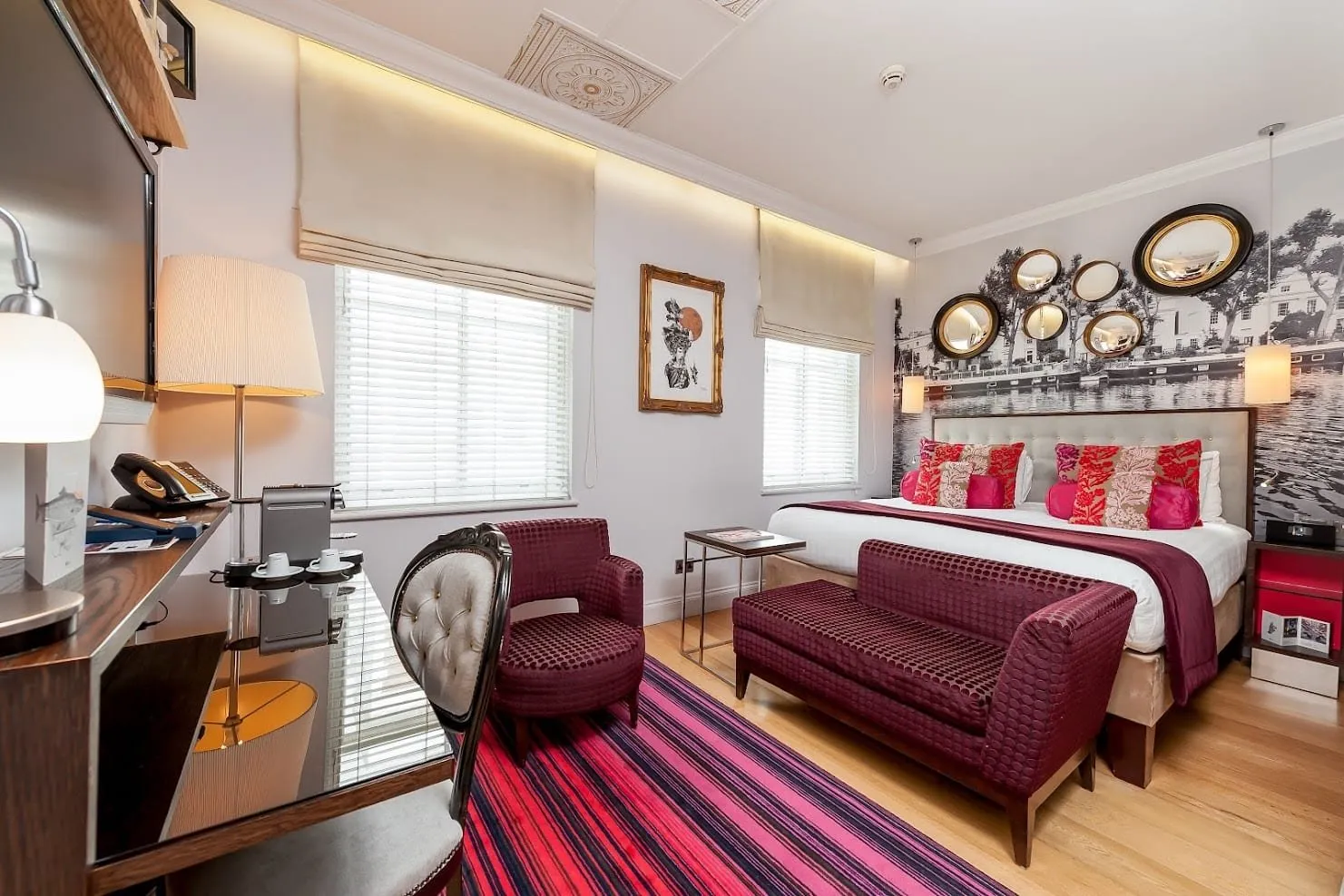 A double room at Hotel Indigo London Paddington with two windows, a black-and-white mural of a canal lined with narrowboats and stucco houses behind the bed, hung with round convex mirrors, burgundy seating and a striped pink rug