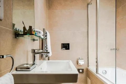 A Hotel Xenia bathroom in sand-coloured tiles with a basin, a glass shelf of toiletries and a bath with a glass shower screen
