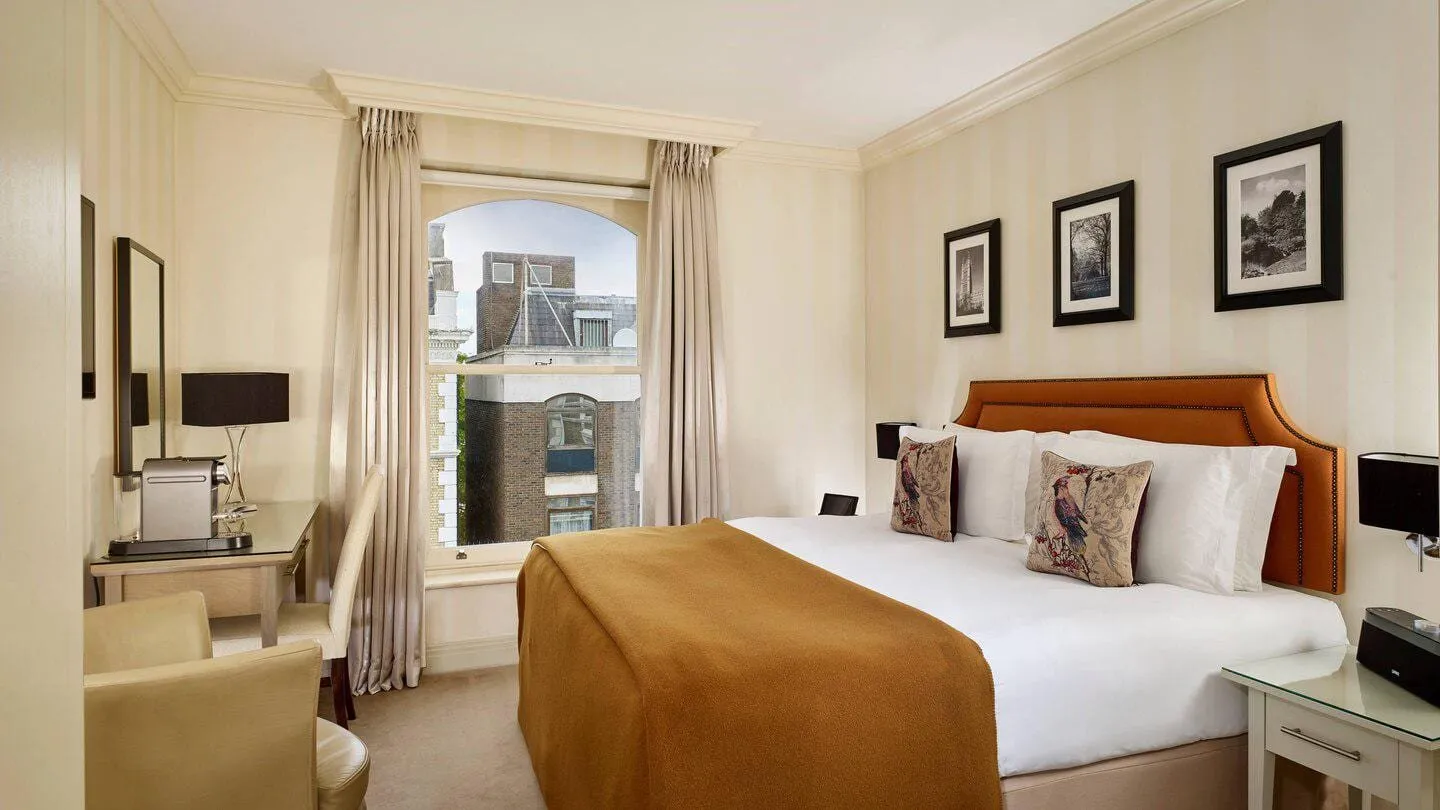 A Hotel Xenia double with a copper studded headboard, bird-print cushions, a caramel throw, a coffee machine on the desk and a tall arched sash window