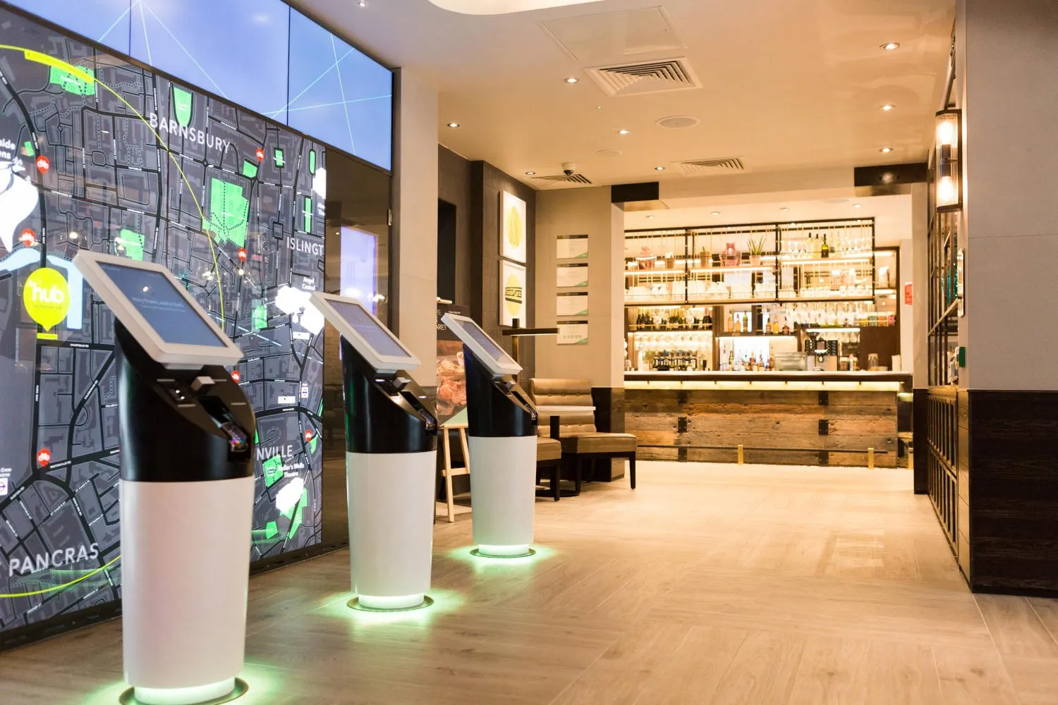Touchscreen check-in kiosks in the lobby of hub by Premier Inn London King's Cross, with the bar behind them