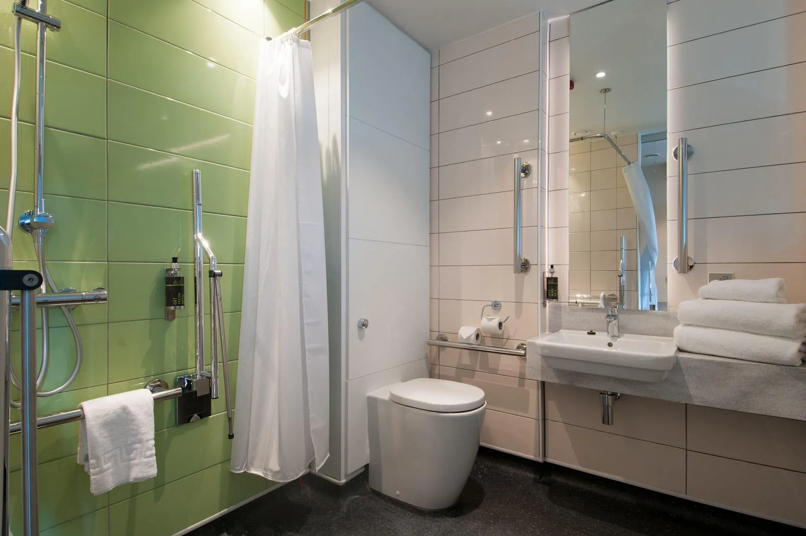 The ensuite bathroom of a hub by Premier Inn Westminster Abbey room, with green tiling, grab rails and a curtained shower