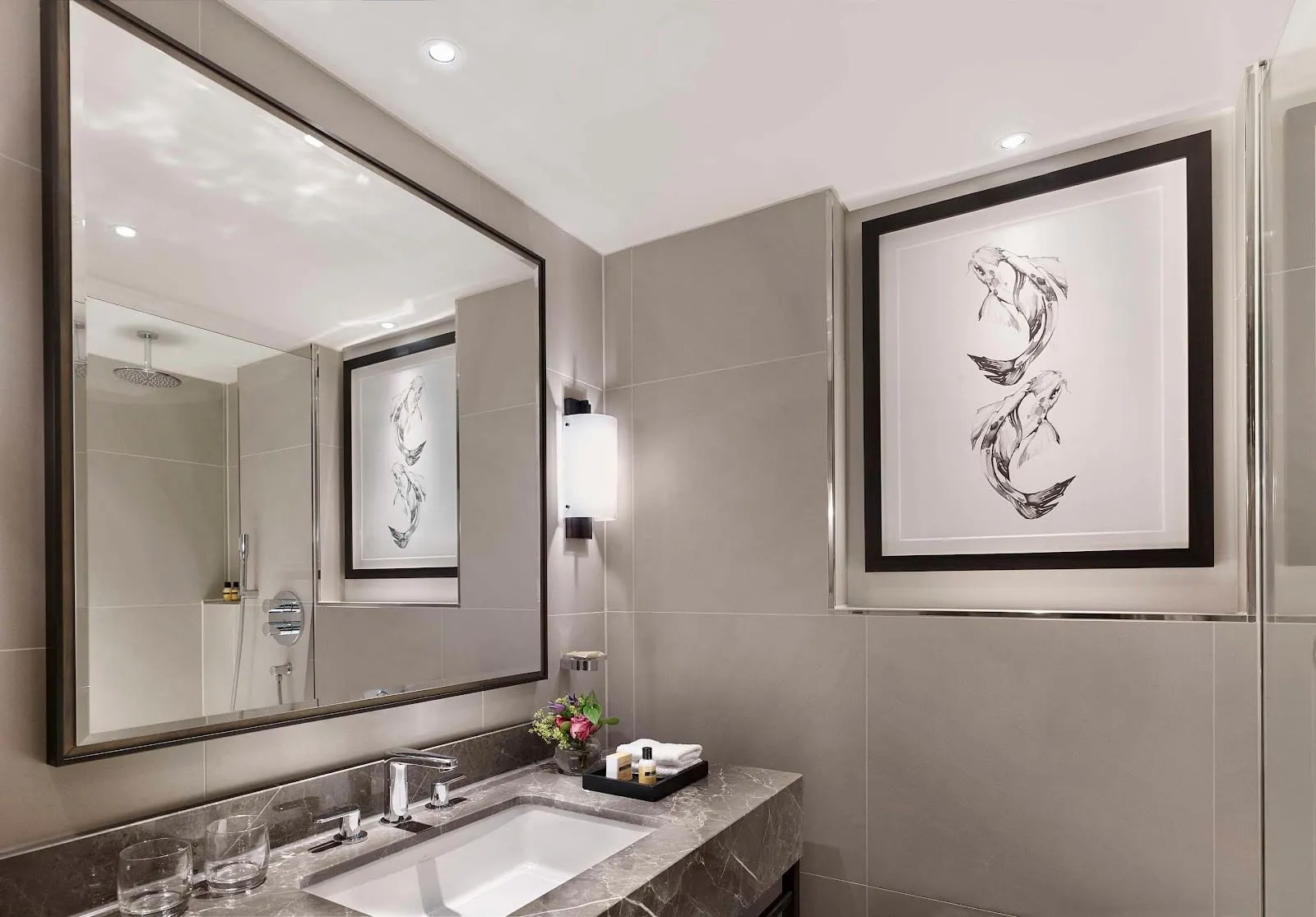 A grey-tiled bathroom at The Churchill with a marble vanity, a framed koi carp drawing, and a walk-in shower with a ceiling rain head reflected in the large mirror