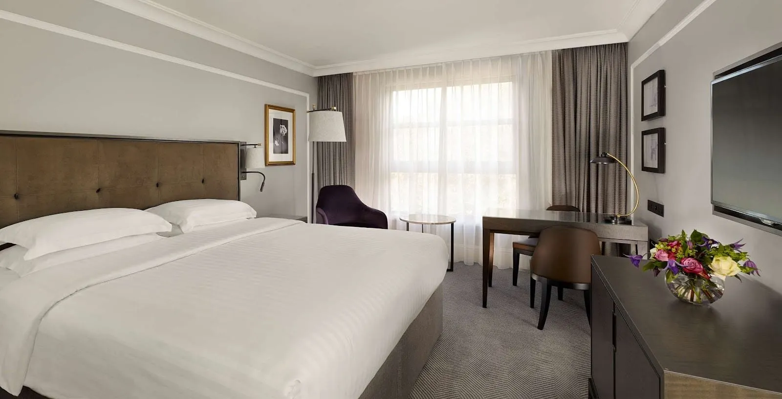 A room at the Hyatt Regency London The Churchill with a king bed against a brown buttoned headboard, grey walls with white panel mouldings, a purple armchair and a desk beside a window with sheer curtains