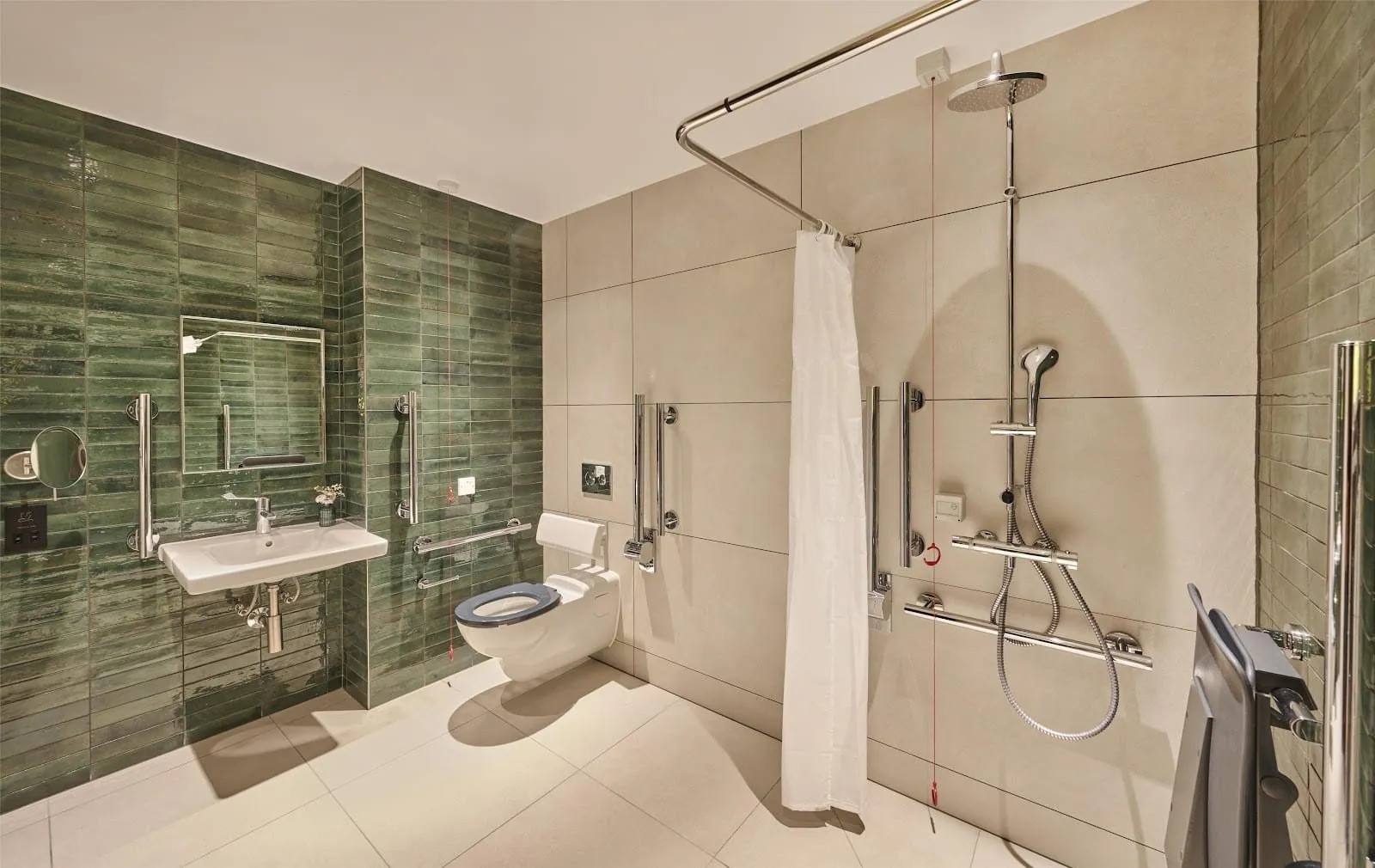 An accessible wet room at the Hyatt Regency London Olympia with green glazed tiles, a level-entry shower with a fold-down seat and grab rails, a toilet with a backrest and an emergency pull cord