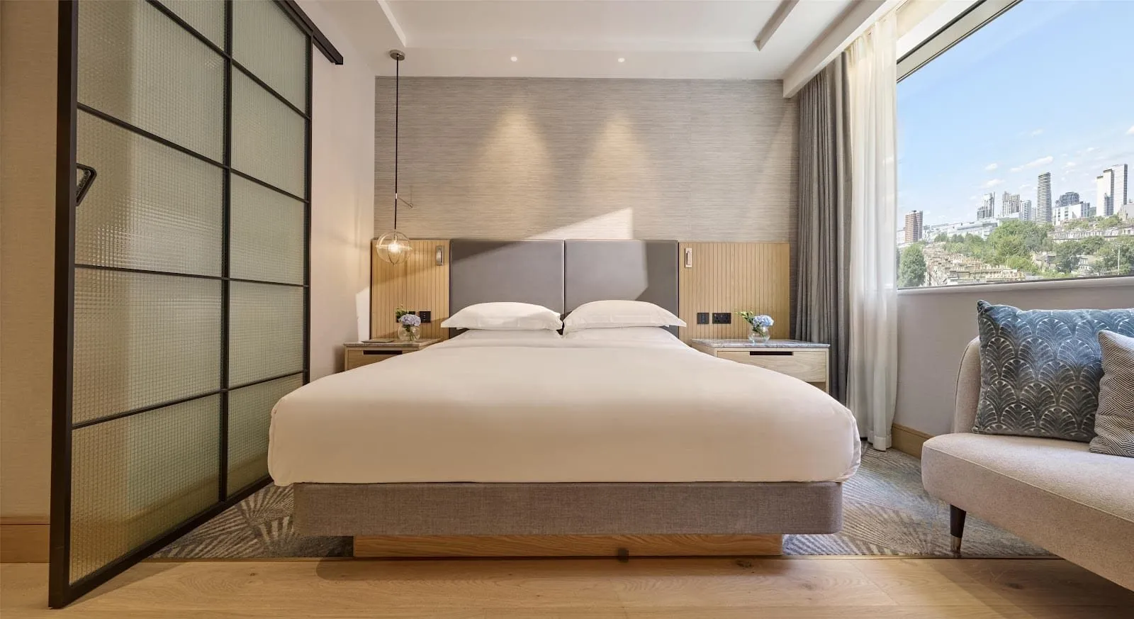 A Hyatt Regency London Olympia room with a king bed on a raised oak platform, a grey headboard, a reeded glass sliding door and a wide window looking over West London terraces to distant towers