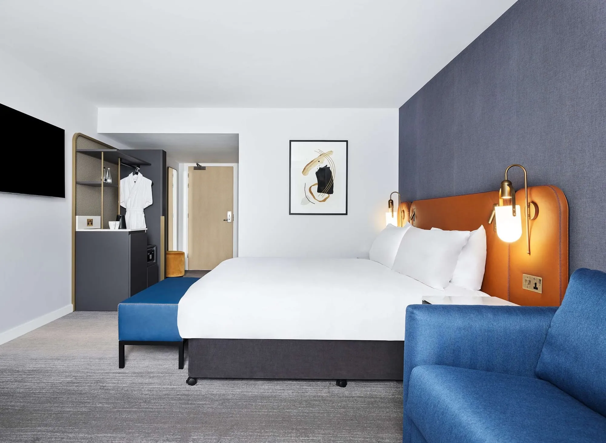 A room at Hyatt Regency London Stratford, with a tan leather headboard, a blue sofa and abstract art on the wall