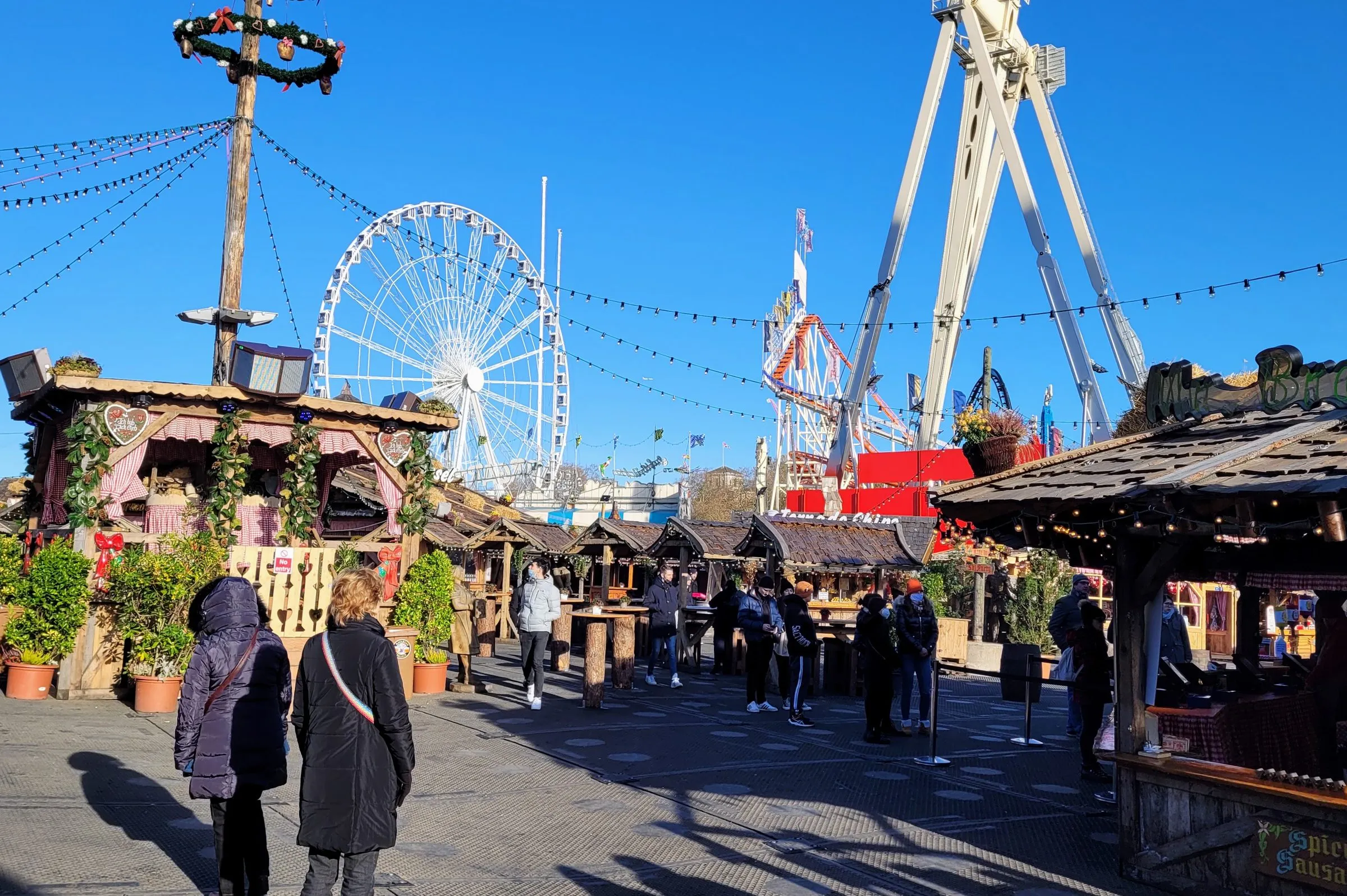 Wooden chalets and a garlanded maypole in Winter Wonderland's Bavarian village on a sunny morning, with the giant wheel and a thrill ride behind and a bratwurst stall on the right