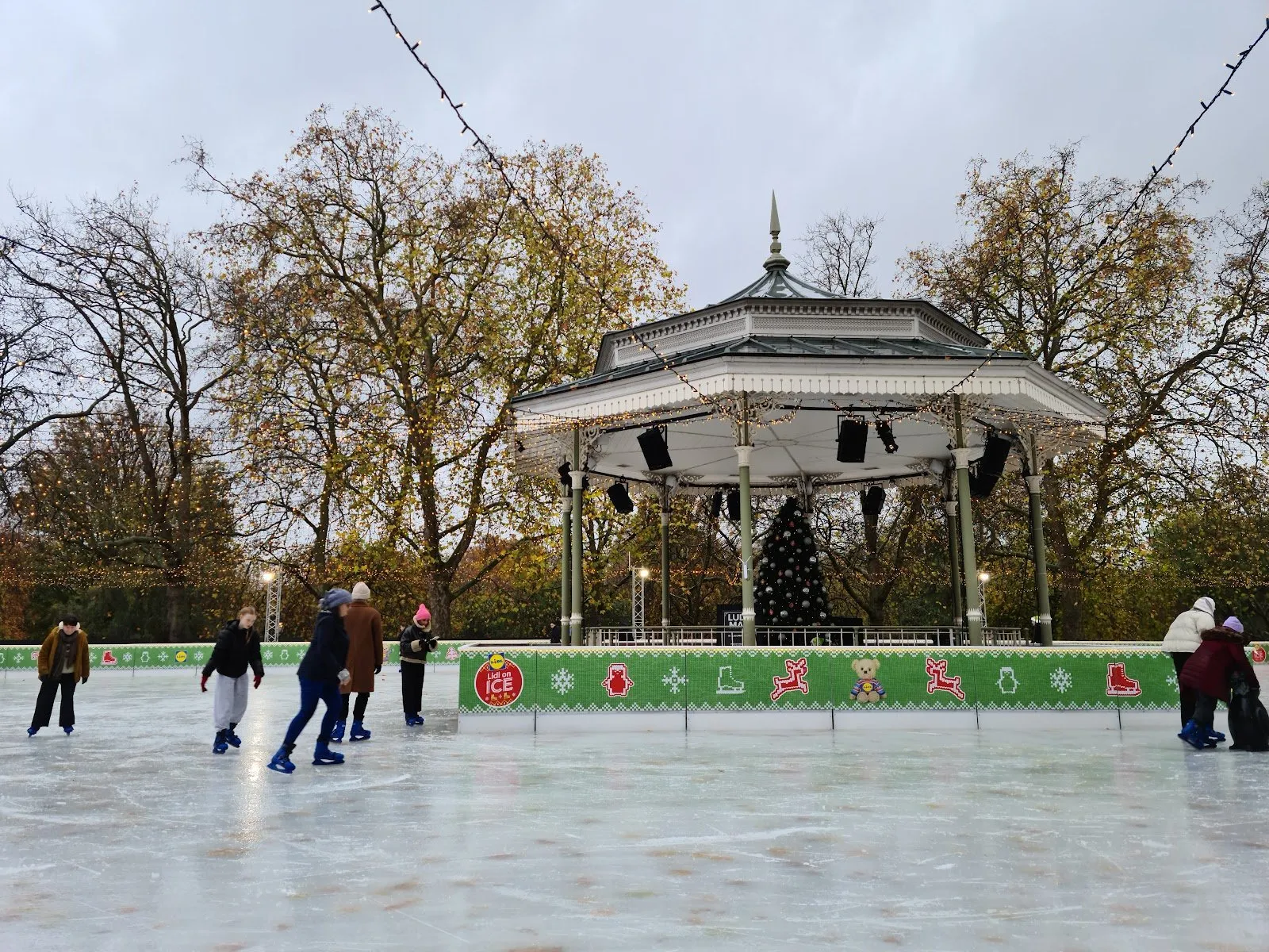 Skaters on the Winter Wonderland ice rink in Hyde Park by day, circling the Victorian bandstand with a Christmas tree inside it