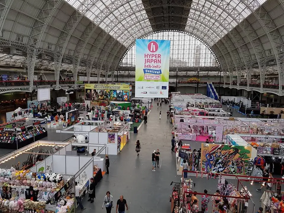 The vaulted glass roof of Olympia's Grand Hall seen from a gallery above, market stalls and a HYPER JAPAN Festival banner filling the hall floor below
