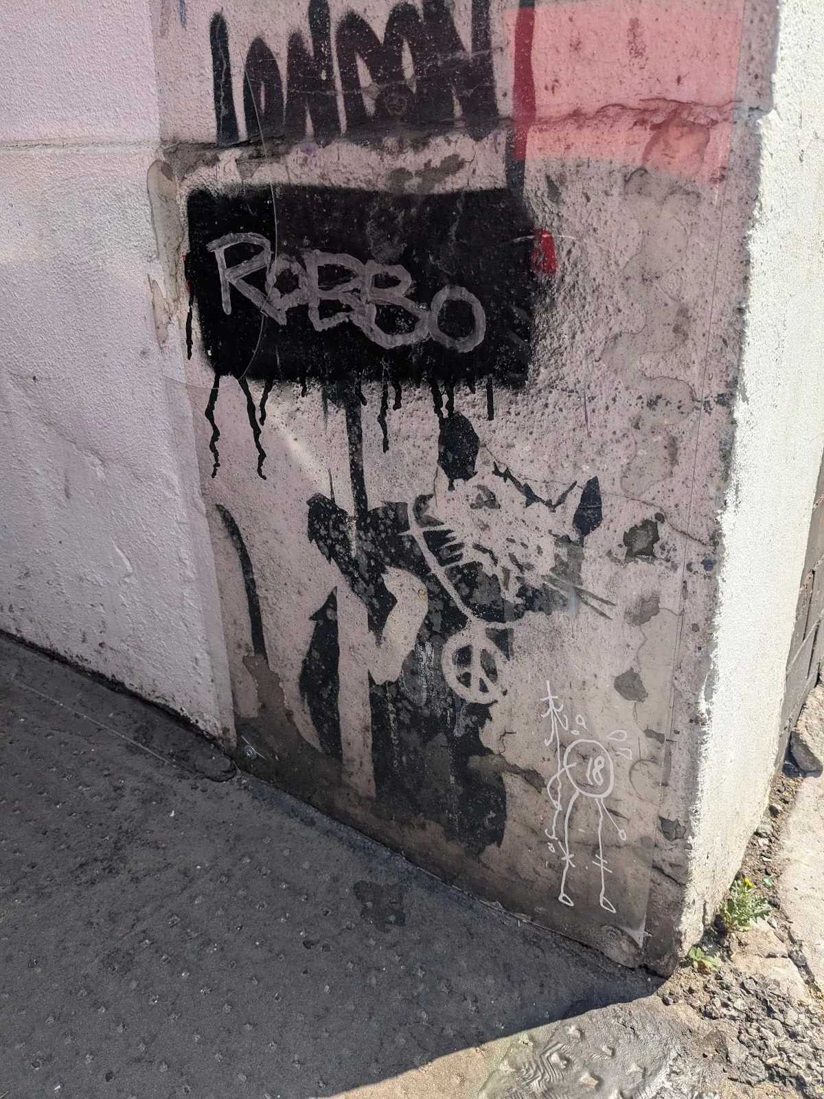 Banksy's rat stencilled low on a white wall corner on Chiswell Street, wearing a peace-sign medallion and holding up a placard reading I love London, with Robbo written in white on a black panel