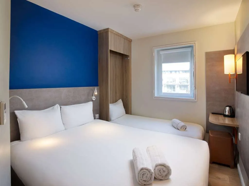 A room at ibis budget London Whitechapel with a double bed, a single bed beside it and a shelf with a kettle