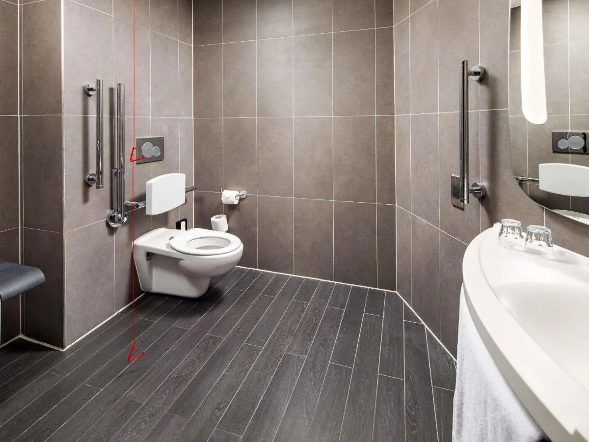 An accessible wet-room bathroom at ibis London Canning Town: grey-brown tiles, a dark wood-effect floor, grab rails, a fold-down seat, a red emergency cord and a curved white basin
