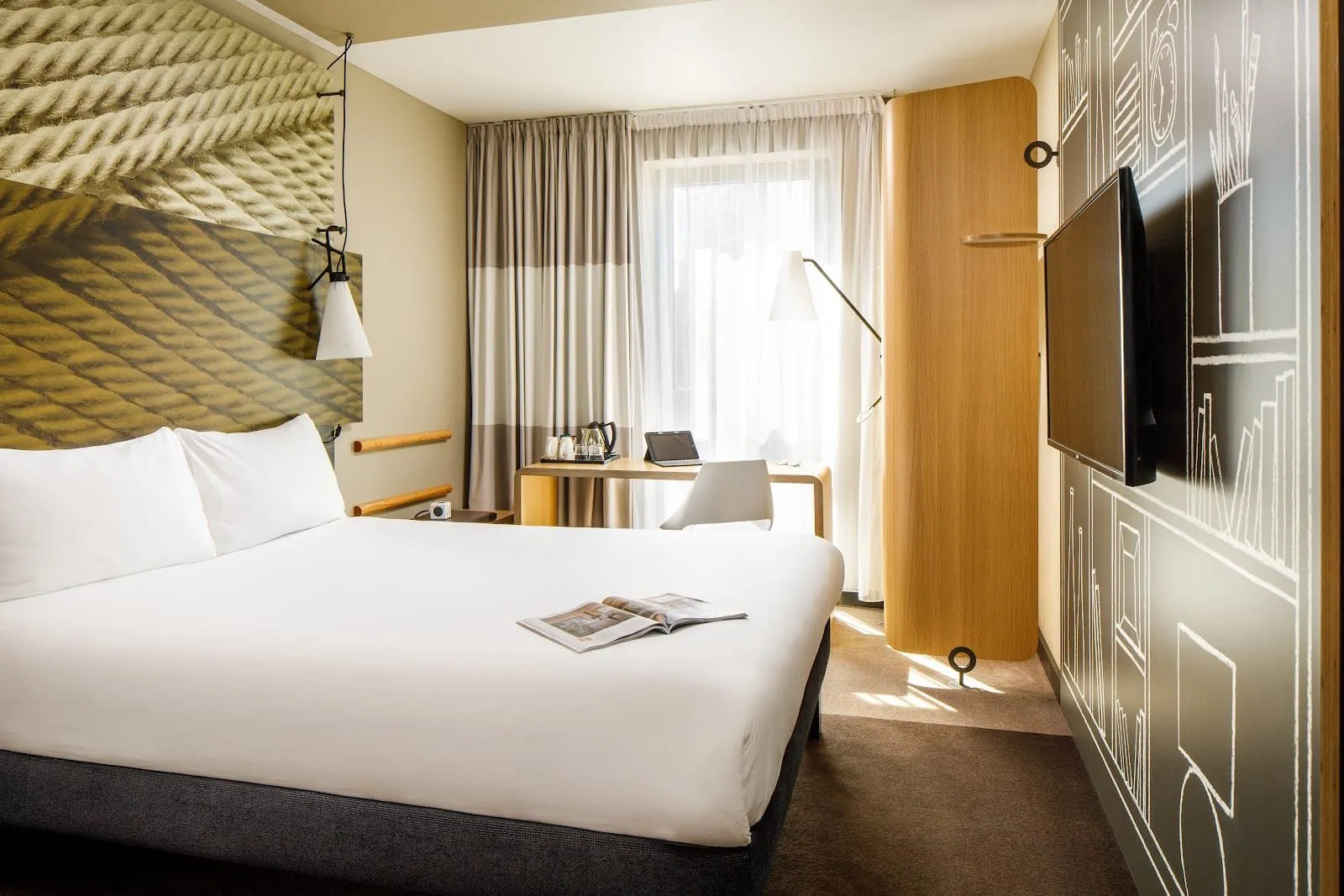 A double room at ibis London Canning Town with a rope-print headboard panel, white bedding, a curved oak wardrobe column, a small desk by a full-height window and a TV on a chalk-sketch wall