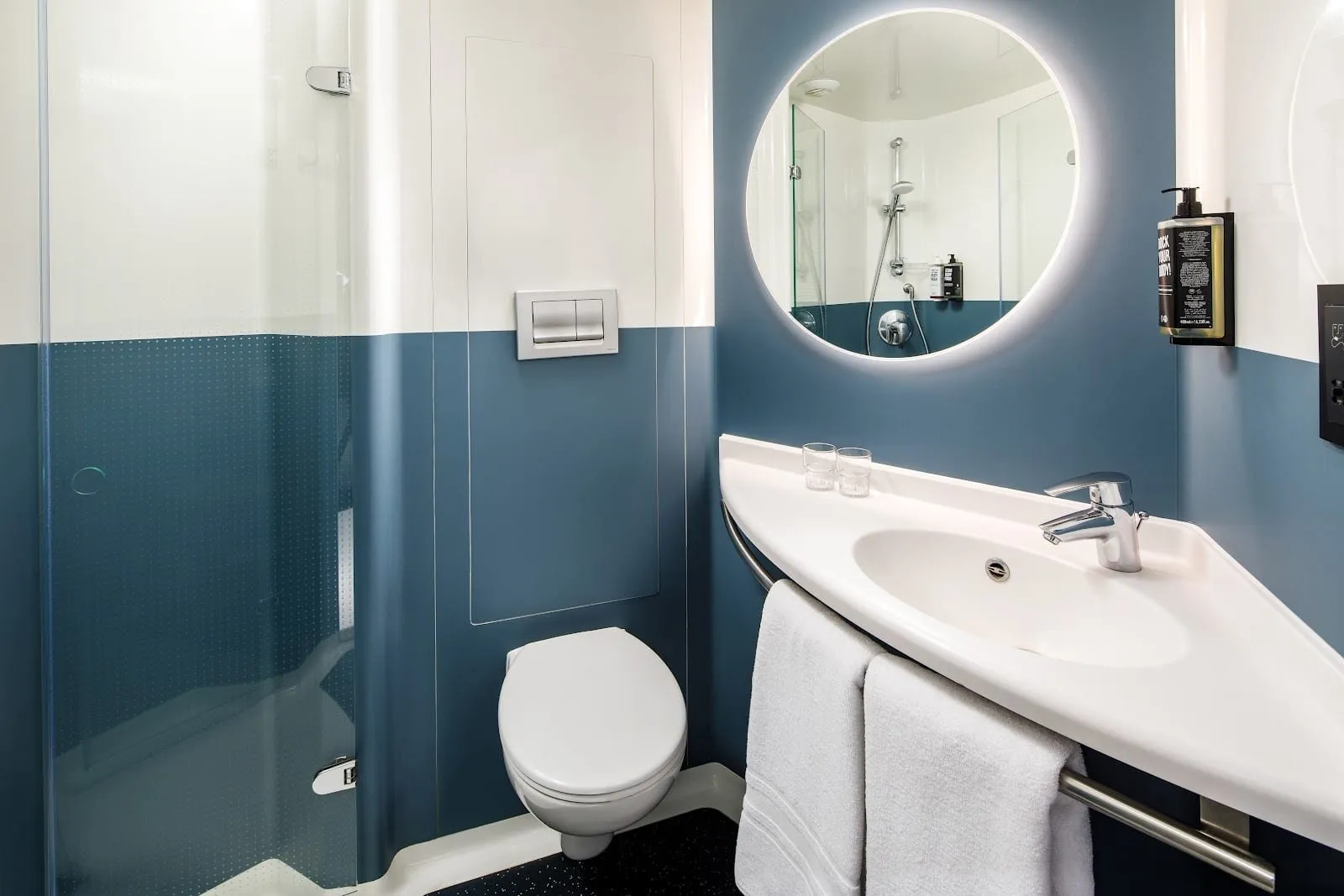 A compact blue-and-white bathroom at ibis London Blackfriars with a walk-in shower, a wall-hung toilet and a corner basin under a round backlit mirror