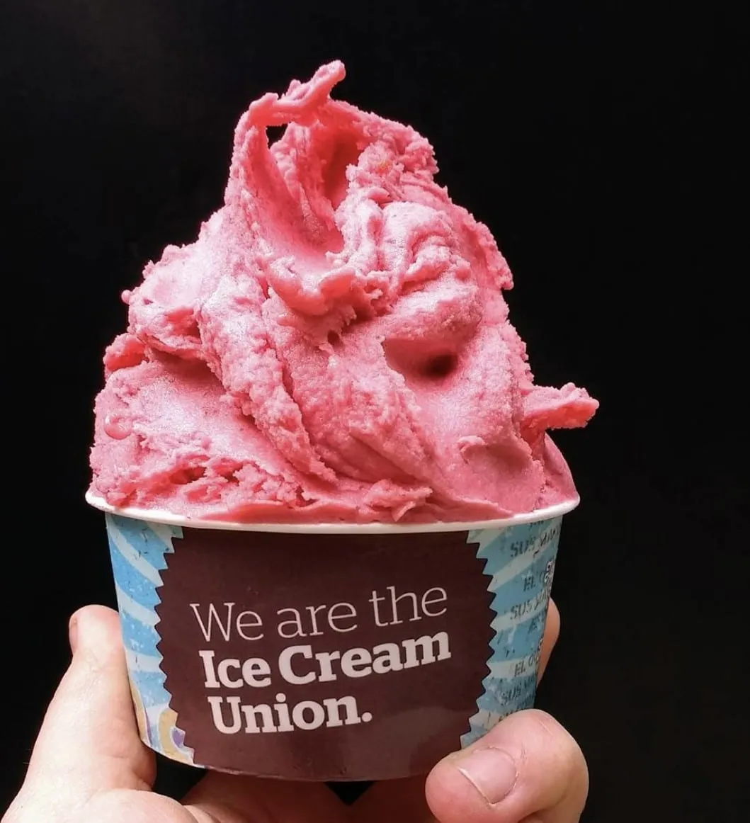 A deep pink fruit sorbet heaped in a tub printed "We are the Ice Cream Union", held against a black background
