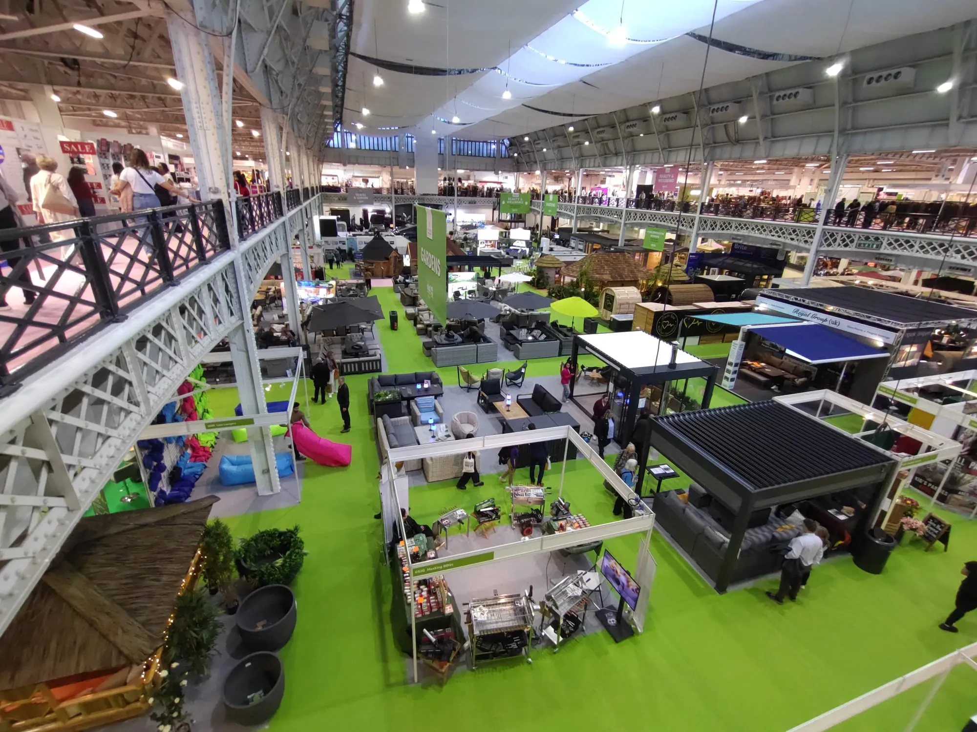 The Ideal Gardens section of the Ideal Home Show, sofas, parasols and garden buildings laid out on a green artificial-turf floor, seen from the mezzanine above