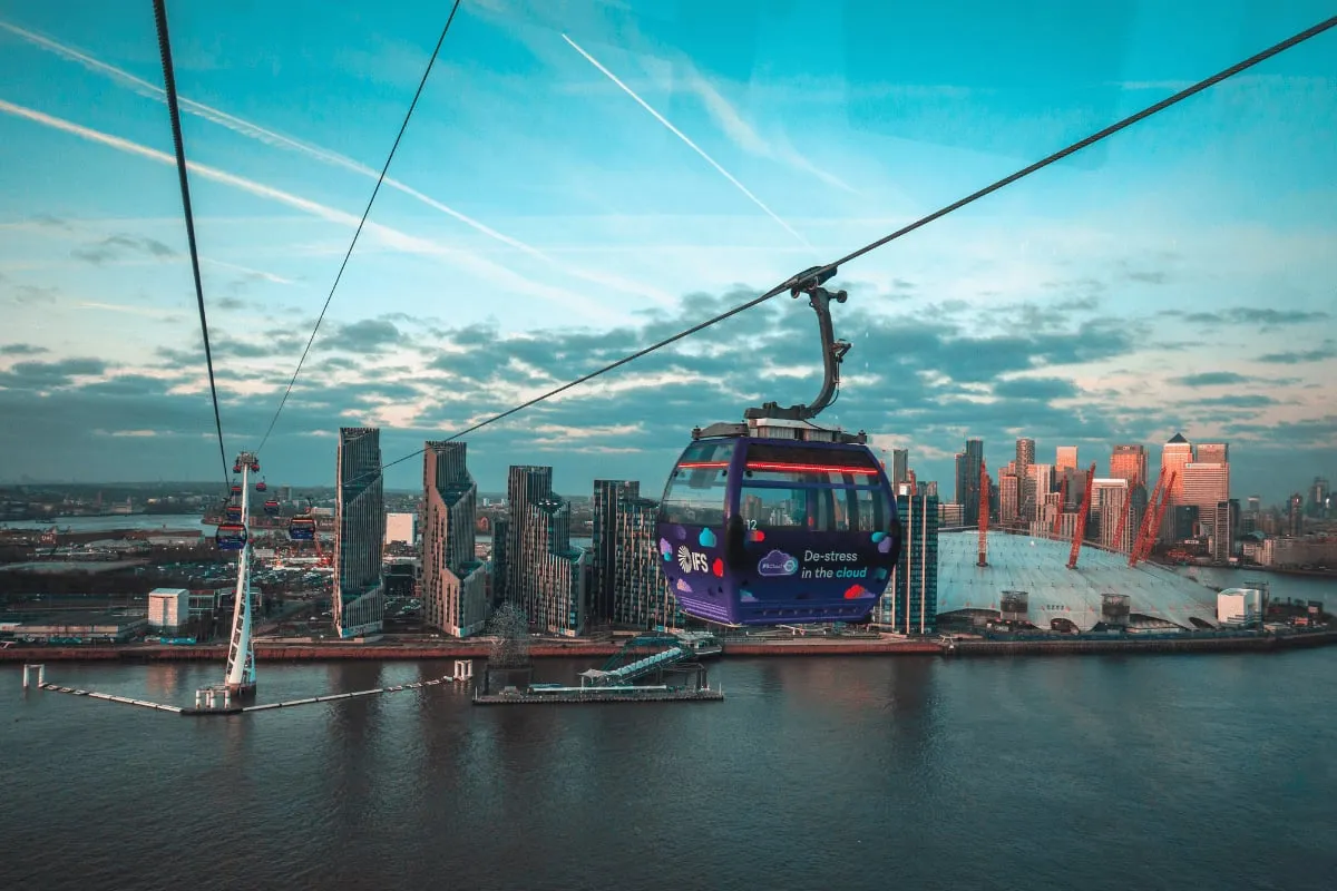 A purple cable car cabin on its cable above the Thames, with riverside tower blocks on the left and the white roof of The O2 on the right at dusk