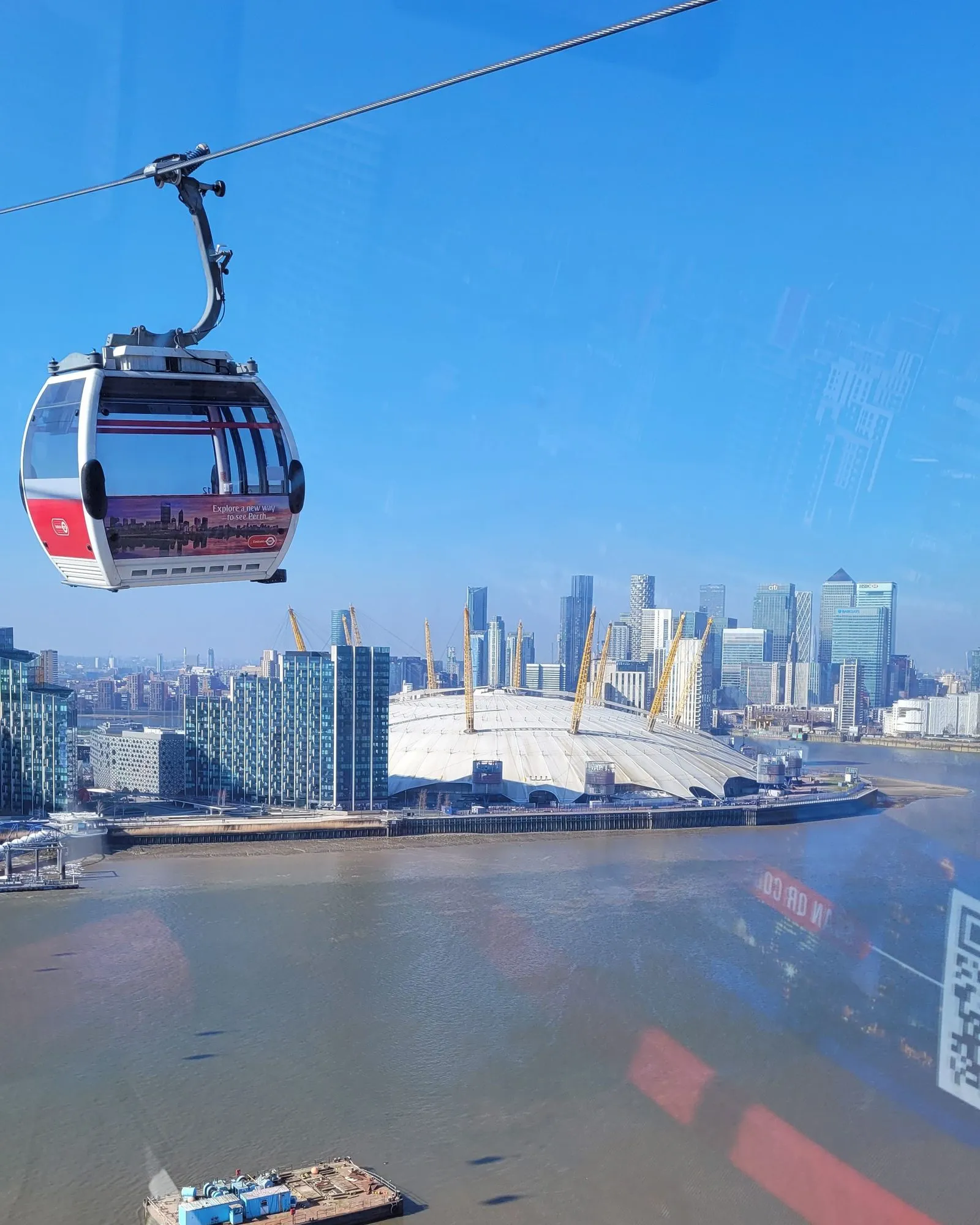 A red and white IFS Cloud Cable Car cabin crossing high above the Thames, with the O2 dome and the Canary Wharf skyline behind