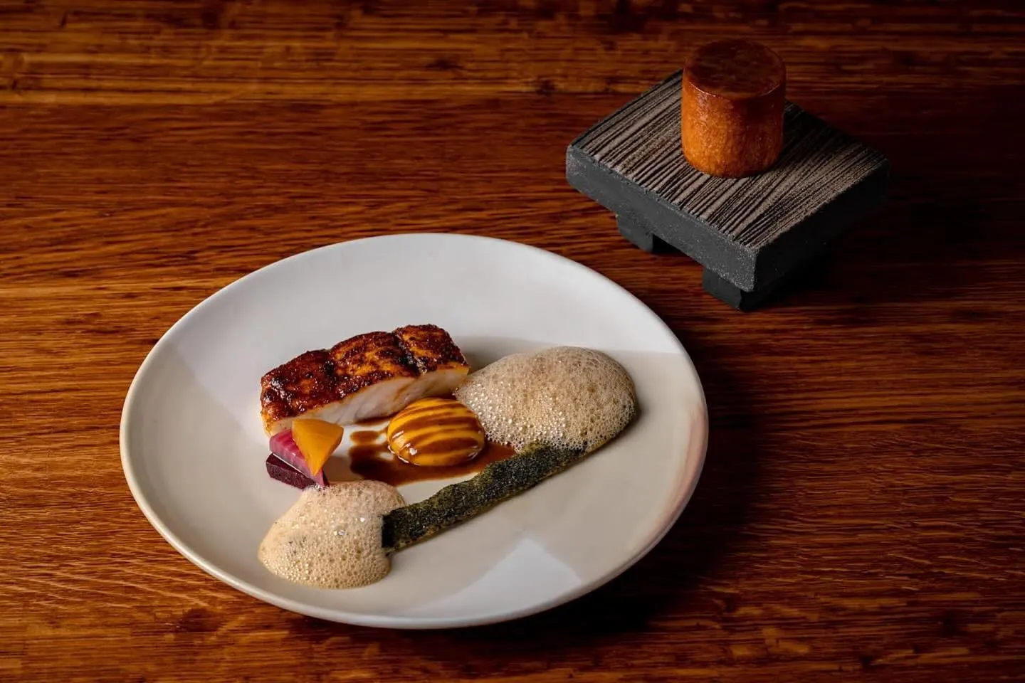 A lacquered fillet glazed almost black at the edges on a pale plate, with a quenelle of orange purée striped in dark sauce, two pools of pale foam, a streak of green oil and a wedge of pickled beetroot; a small cylindrical loaf sits on a ridged black stand beside it