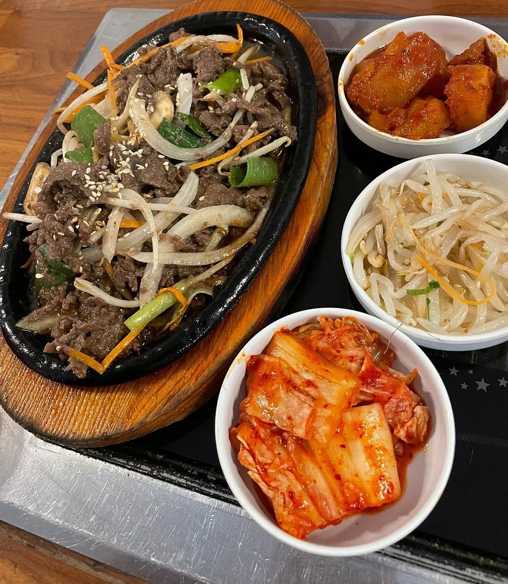 A cast-iron platter of sizzling beef bulgogi with onion, spring onion and sesame at Imone, with bowls of cabbage kimchi, seasoned bean sprouts and cubed radish kimchi beside it