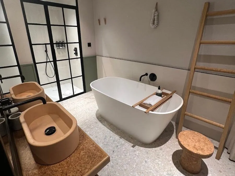 A bathroom in one of the larger rooms at Inhabit, Queen's Gardens with a terrazzo floor, a freestanding white bath, two terracotta basins on a cork counter, a wooden ladder towel rail and a walk-in shower behind black-framed glass