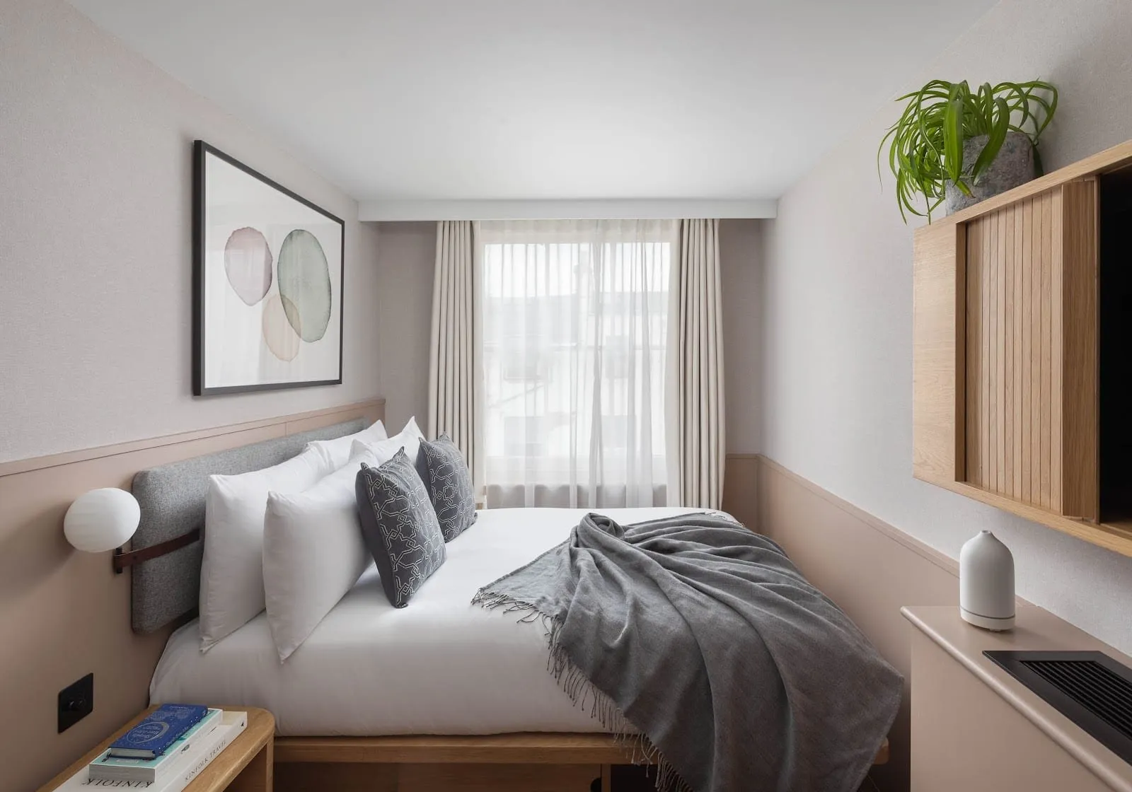 A small double at Inhabit, Queen's Gardens with pale pink walls, a grey fabric headboard, a grey throw, a watercolour print above the bed and an oak cabinet on the wall, the bed filling the width of the room up to the window