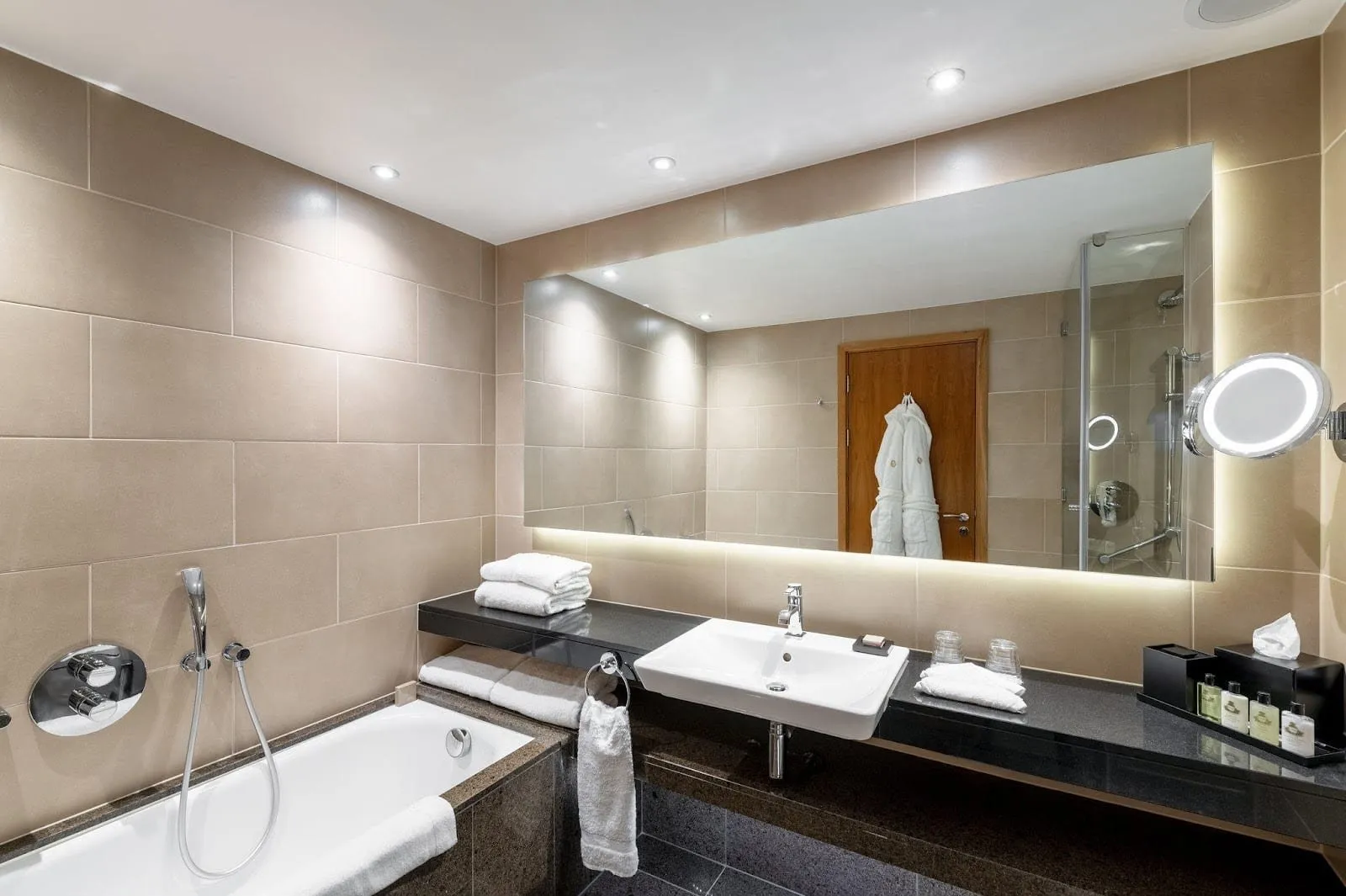The bathroom at InterContinental London – The O2: a tiled bath with a hand shower, a basin on a black granite counter under a long backlit mirror, and a separate glass shower reflected behind