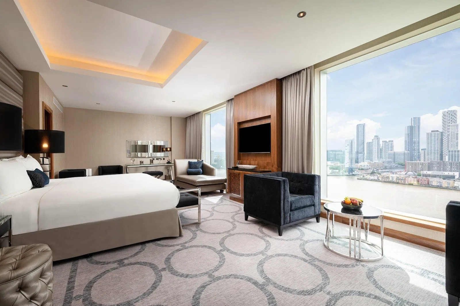A large room at InterContinental London – The O2 with a king bed, a velvet armchair and chaise, a walnut TV wall and a picture window looking across the Thames to the Canary Wharf towers