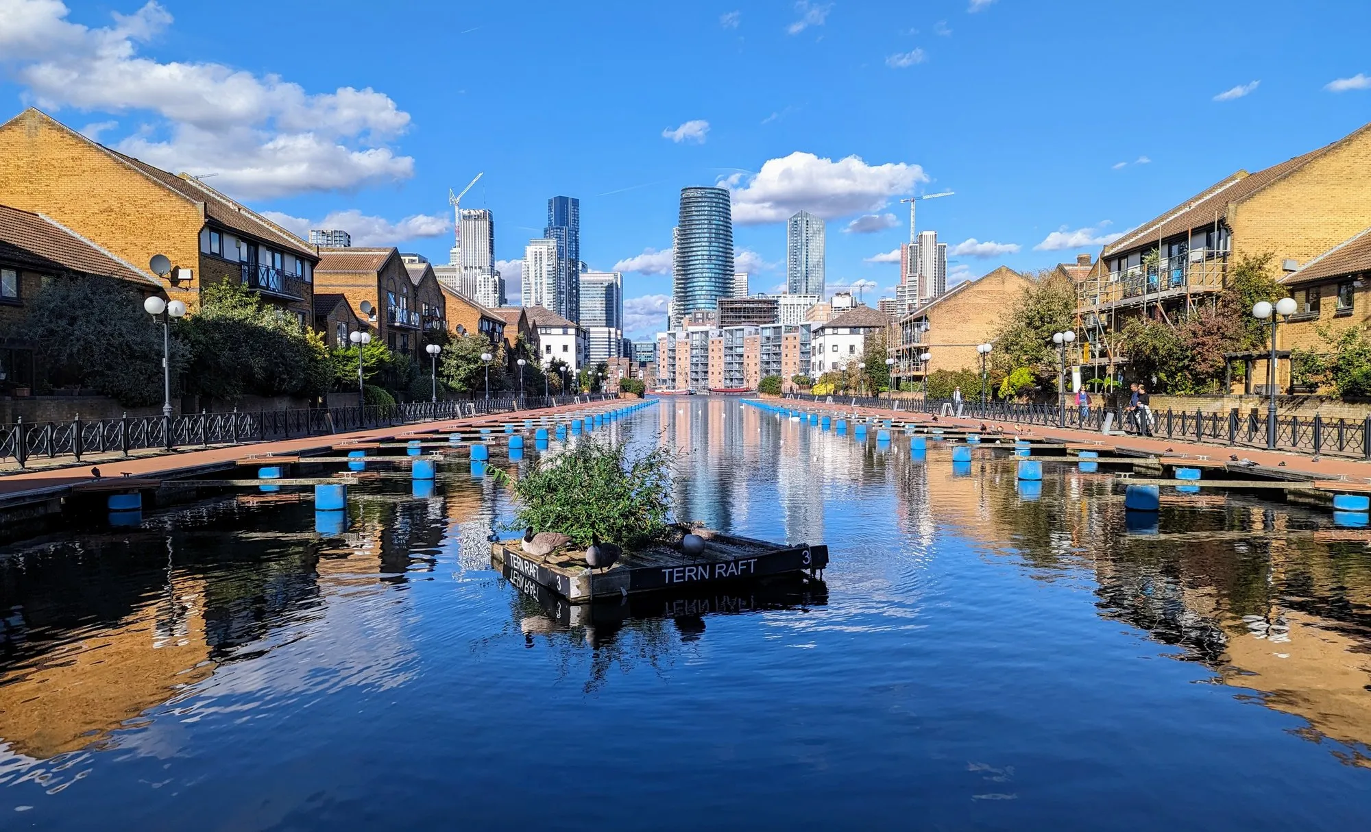 A canal-side dock on the Isle of Dogs lined with converted warehouse flats, with the Canary Wharf skyline reflected in the water and a floating "Tern Raft" nesting platform in the foreground