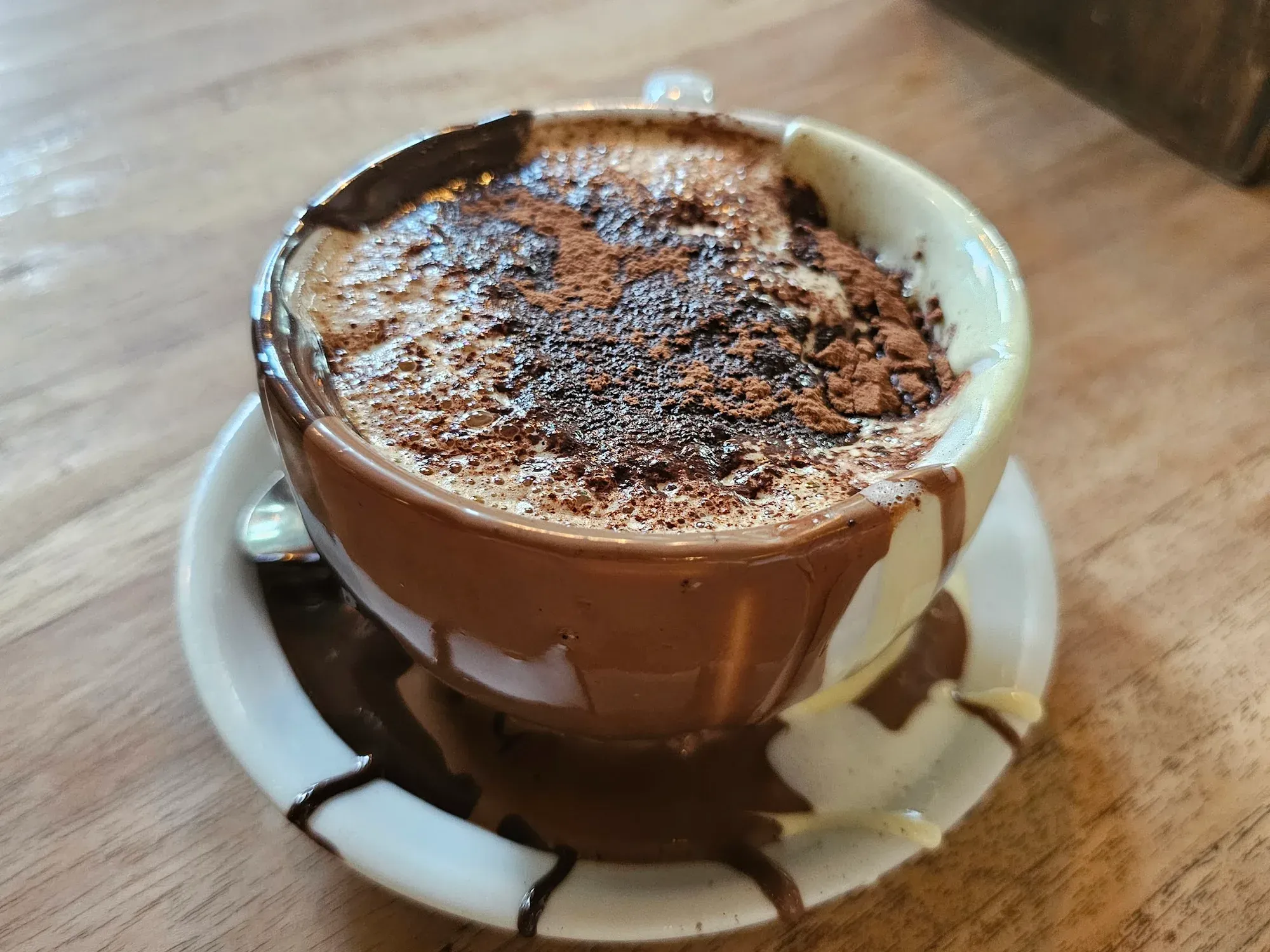 A cup of thick Italian hot chocolate dusted with cocoa, the cup lined with melted chocolate that has run down the outside and pooled on the saucer
