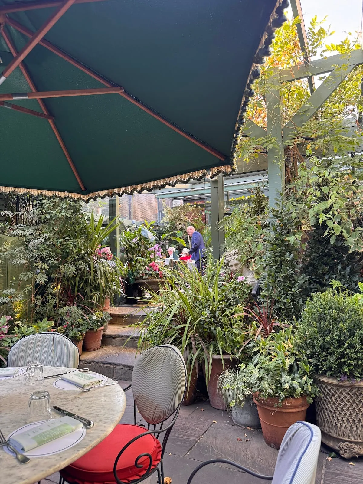 The walled garden at The Ivy Chelsea Garden, café tables under a green scalloped umbrella surrounded by potted plants and climbers