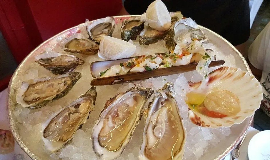 A silver platter on crushed ice holding a dozen oysters, a razor clam, a dressed scallop in its shell and lemon wedges