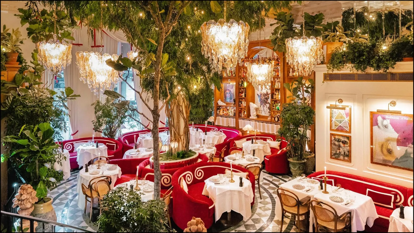 The dining room at Jacuzzi, red curved booths and white tables around indoor trees, under Murano glass chandeliers hung with fairy lights
